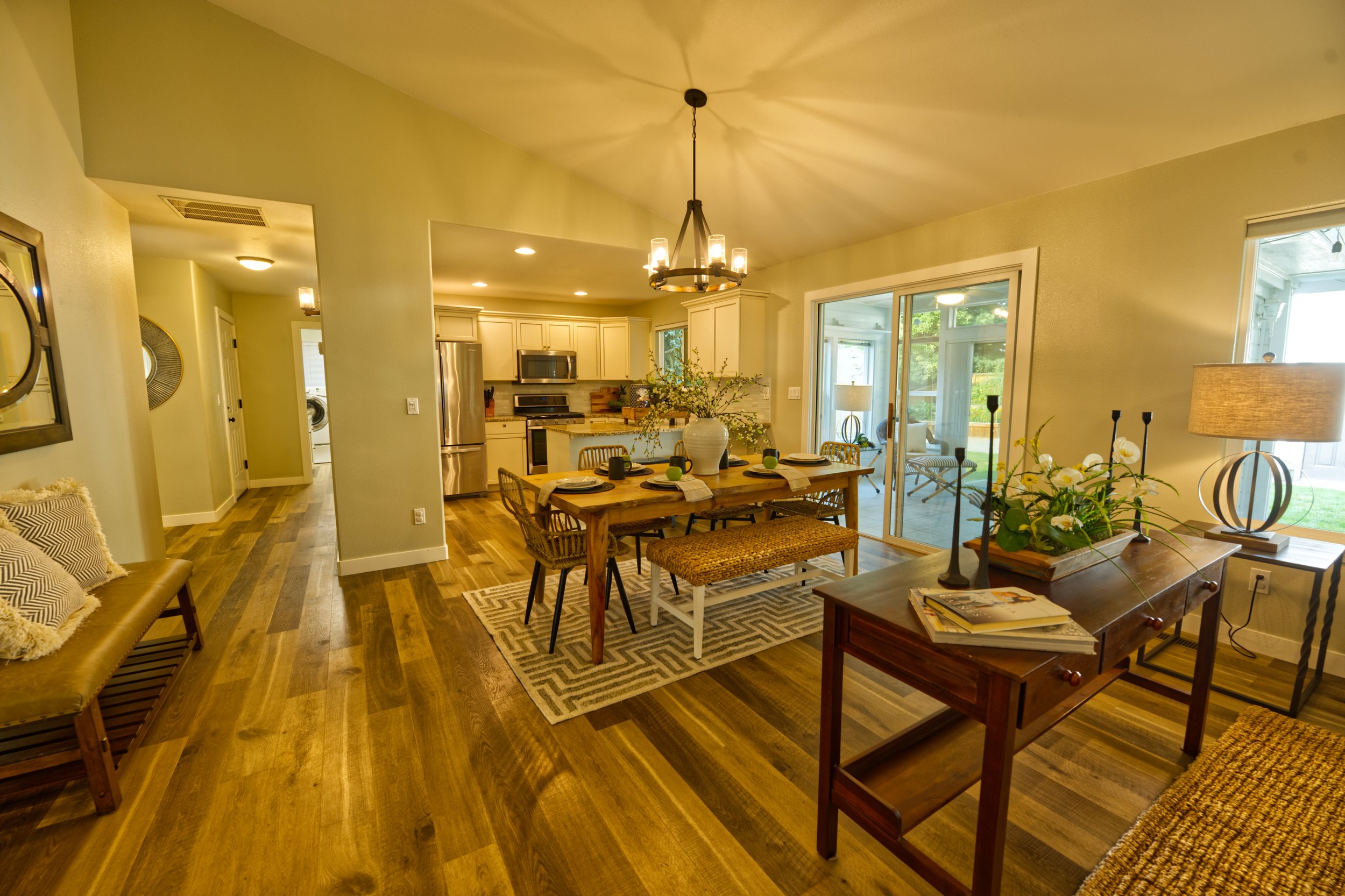 Open-concept living and dining area with wood flooring, a wooden dining table with chairs, a sofa with pillows, and kitchen in the background. Large windows and sliding glass doors let in natural light, with views of greenery outside.