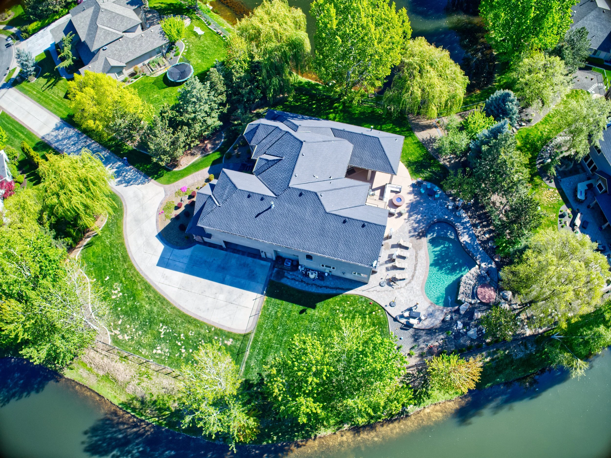 Aerial view of a house surrounded by lush green trees, a well-maintained lawn, and a swimming pool, with a waterway bordering the property.