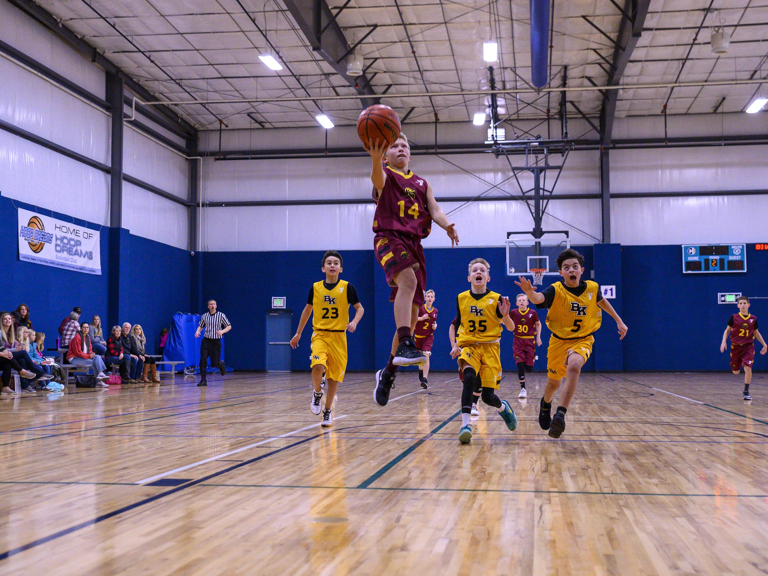 Young boys playing basketball in an indoor gymnasium. One boy in a maroon jersey with the number 14 is jumping to shoot or dunk the ball, while others in yellow jerseys with numbers 23, 35, and 5 are on the court. Spectators are seated along the side