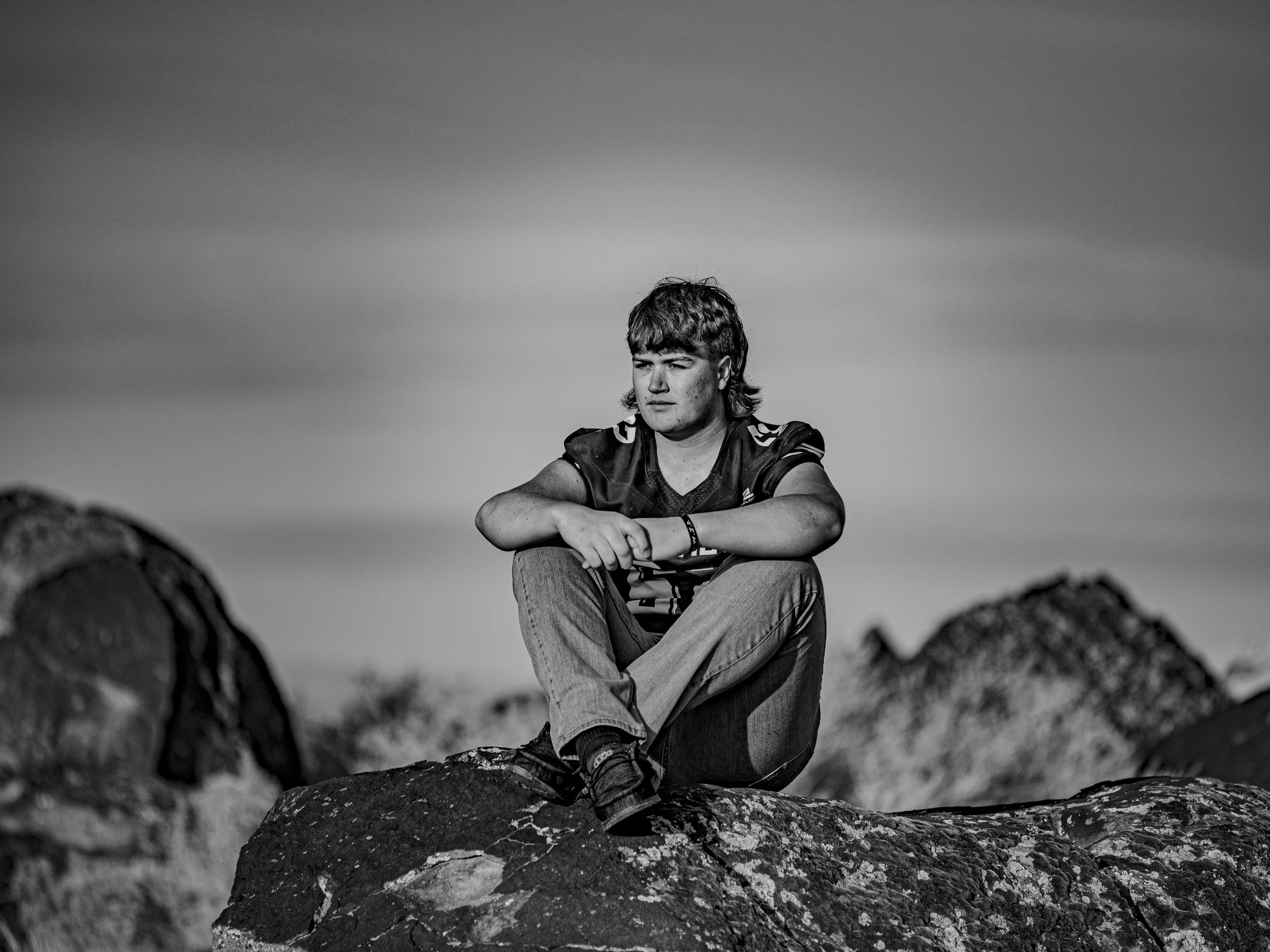 Black and white photo of a young man sitting on a rock outdoors, with short hair and a serious expression, wearing a sports shirt, looking into the distance. Background shows a cloudy sky.