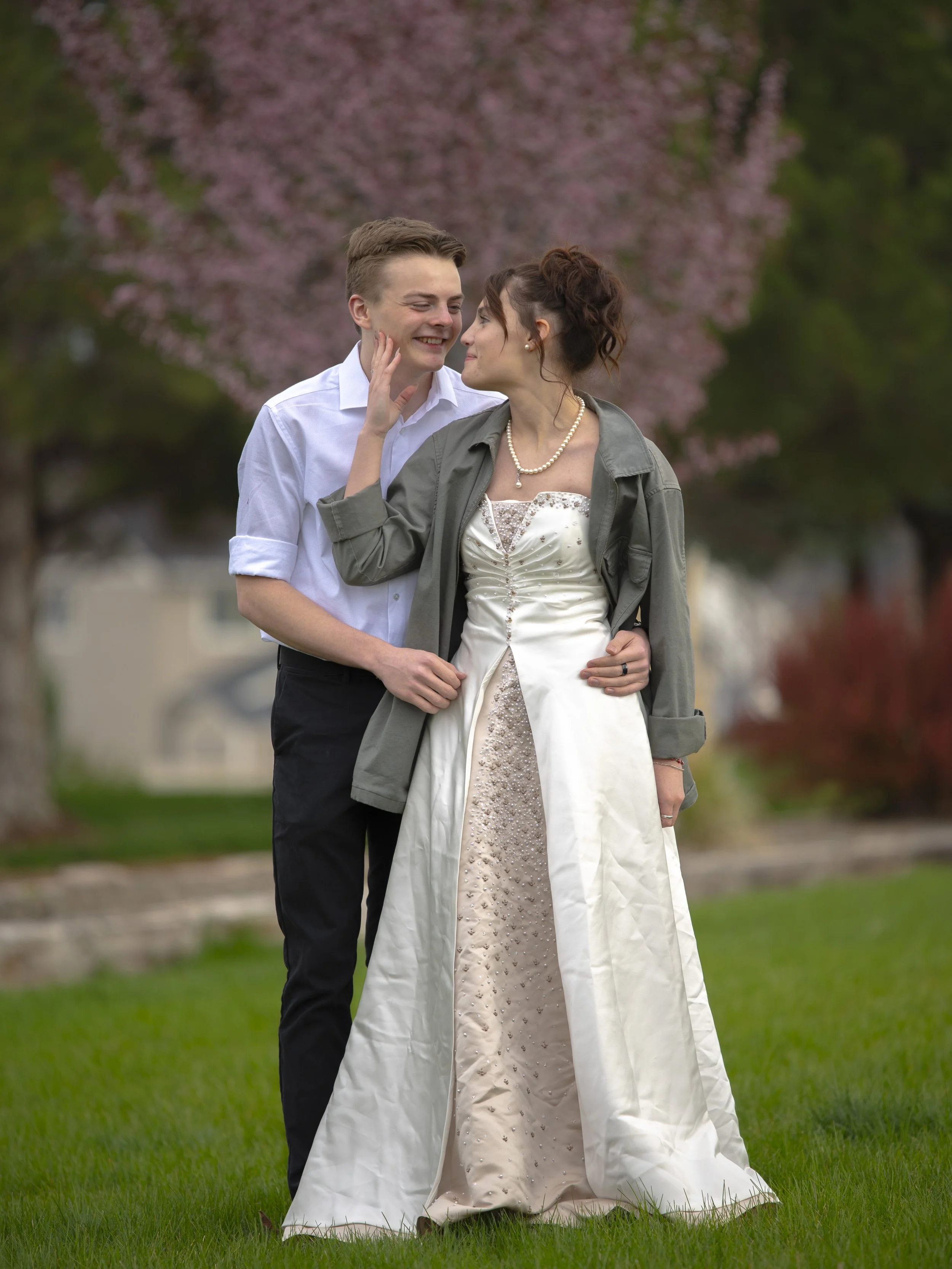 A young couple dressed in wedding attire standing on a grassy lawn with pink flowering trees in the background, sharing an intimate moment.
