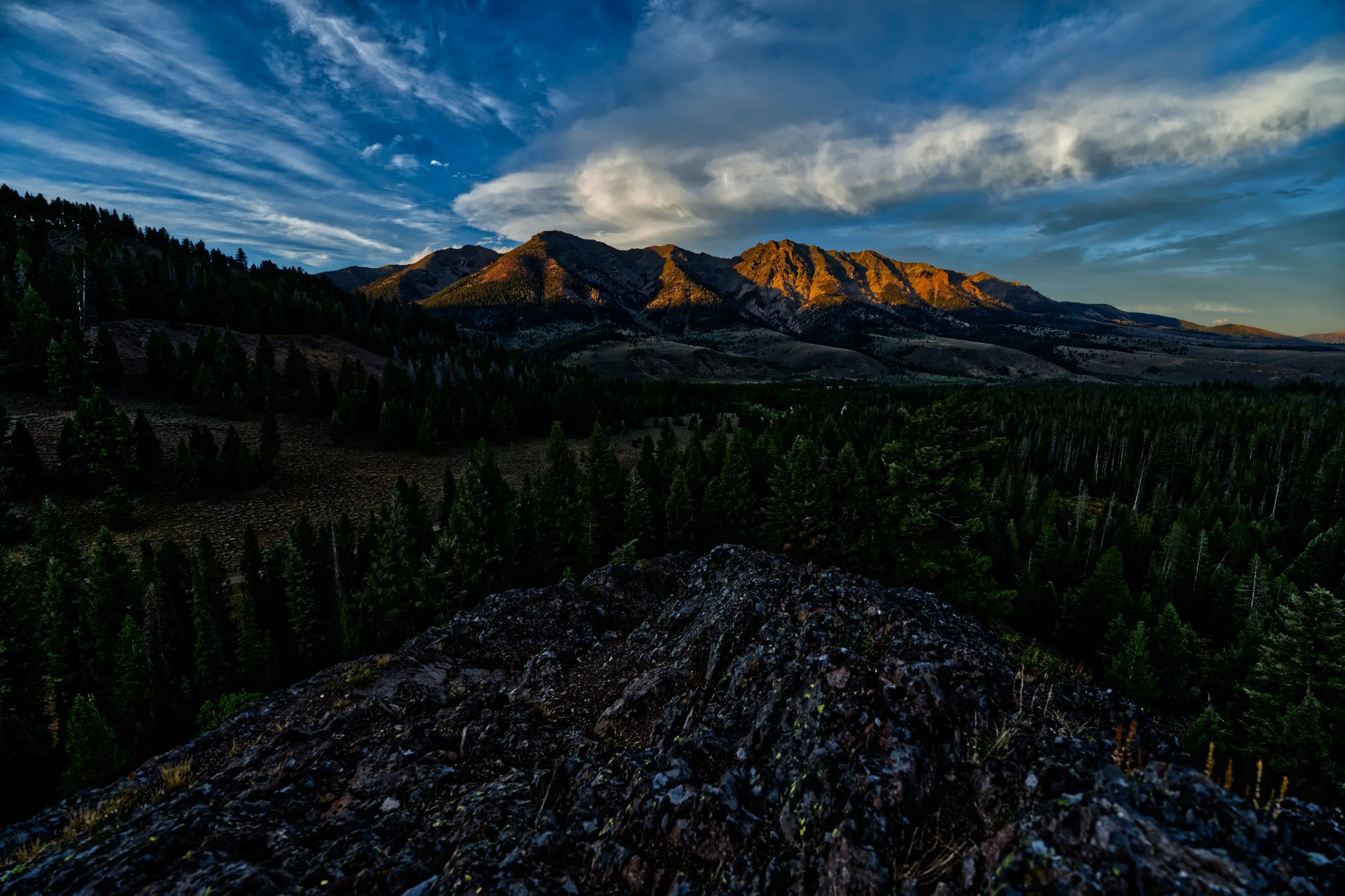 A scenic view of a mountain range during sunset with the peaks illuminated in golden light, a forested valley below, and a dramatic sky with scattered clouds.