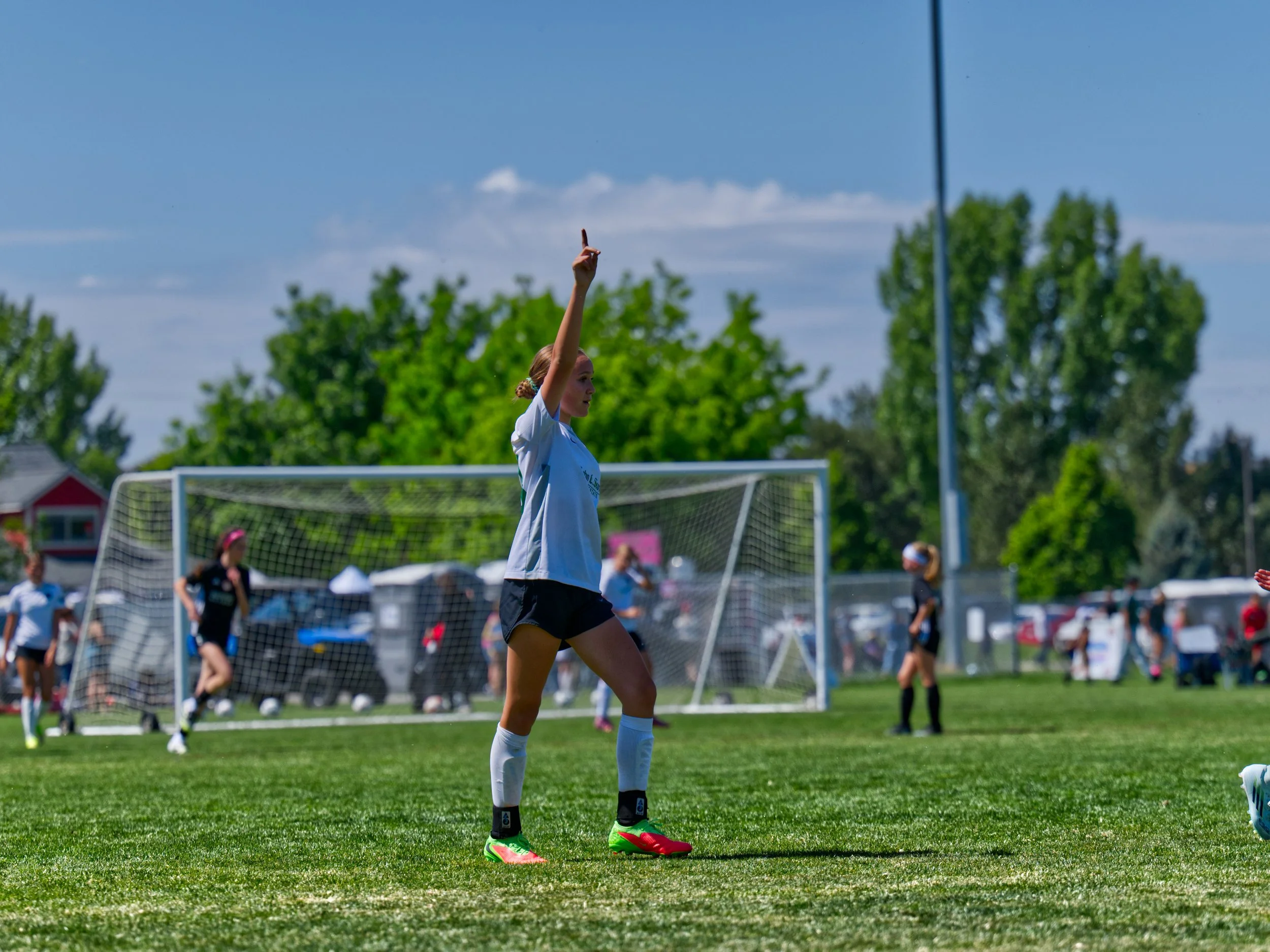 A female soccer player in a white jersey and black shorts on a soccer field, preparing for a game or practice, with a goalpost and other players in the background on a sunny day.