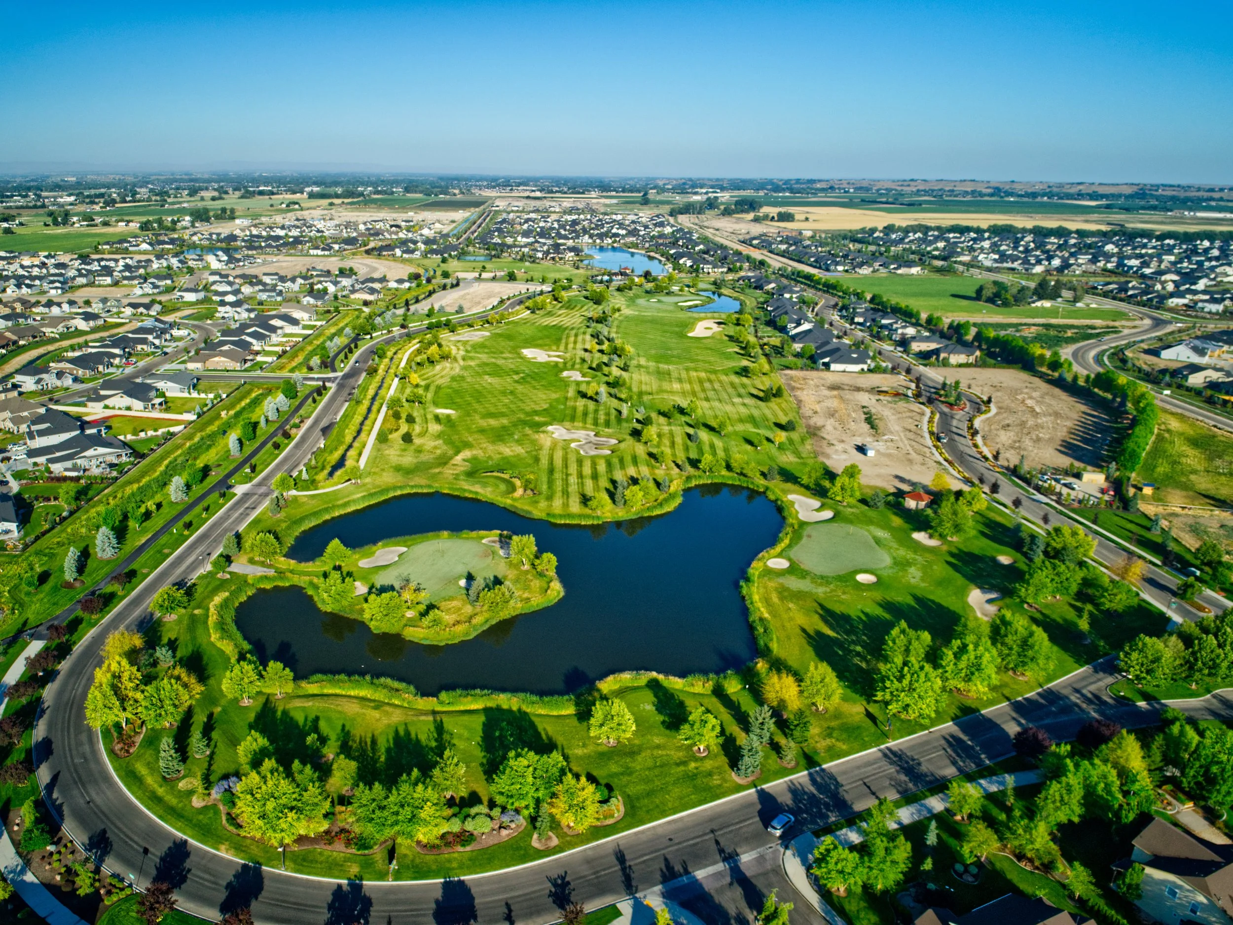 Aerial view of a golf course surrounded by a residential neighborhood with houses, trees, water features, and a park-like setting under a clear blue sky.