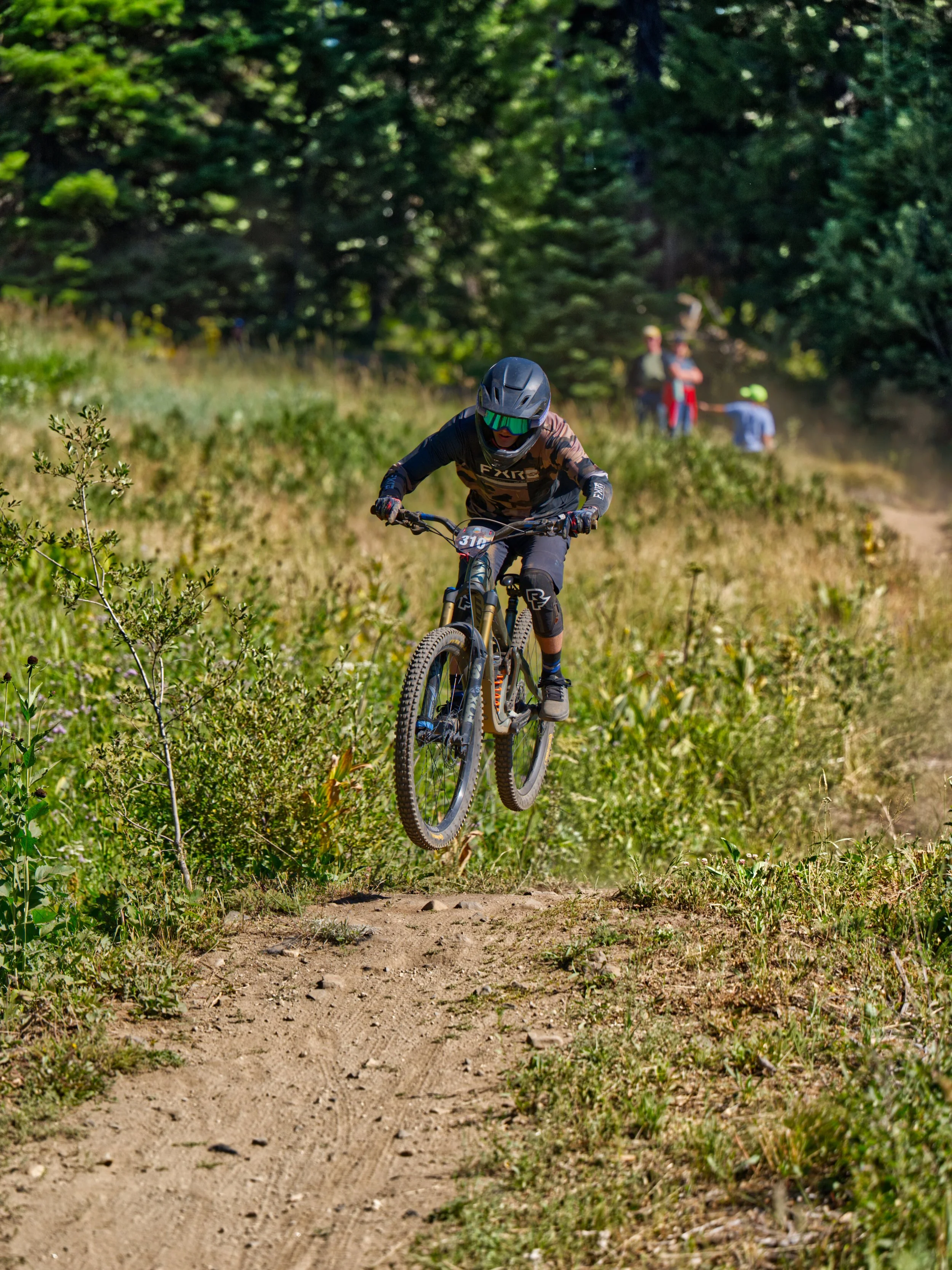 A mountain biker wearing a helmet and goggles riding on a dirt trail through a forest, with spectators in the background.