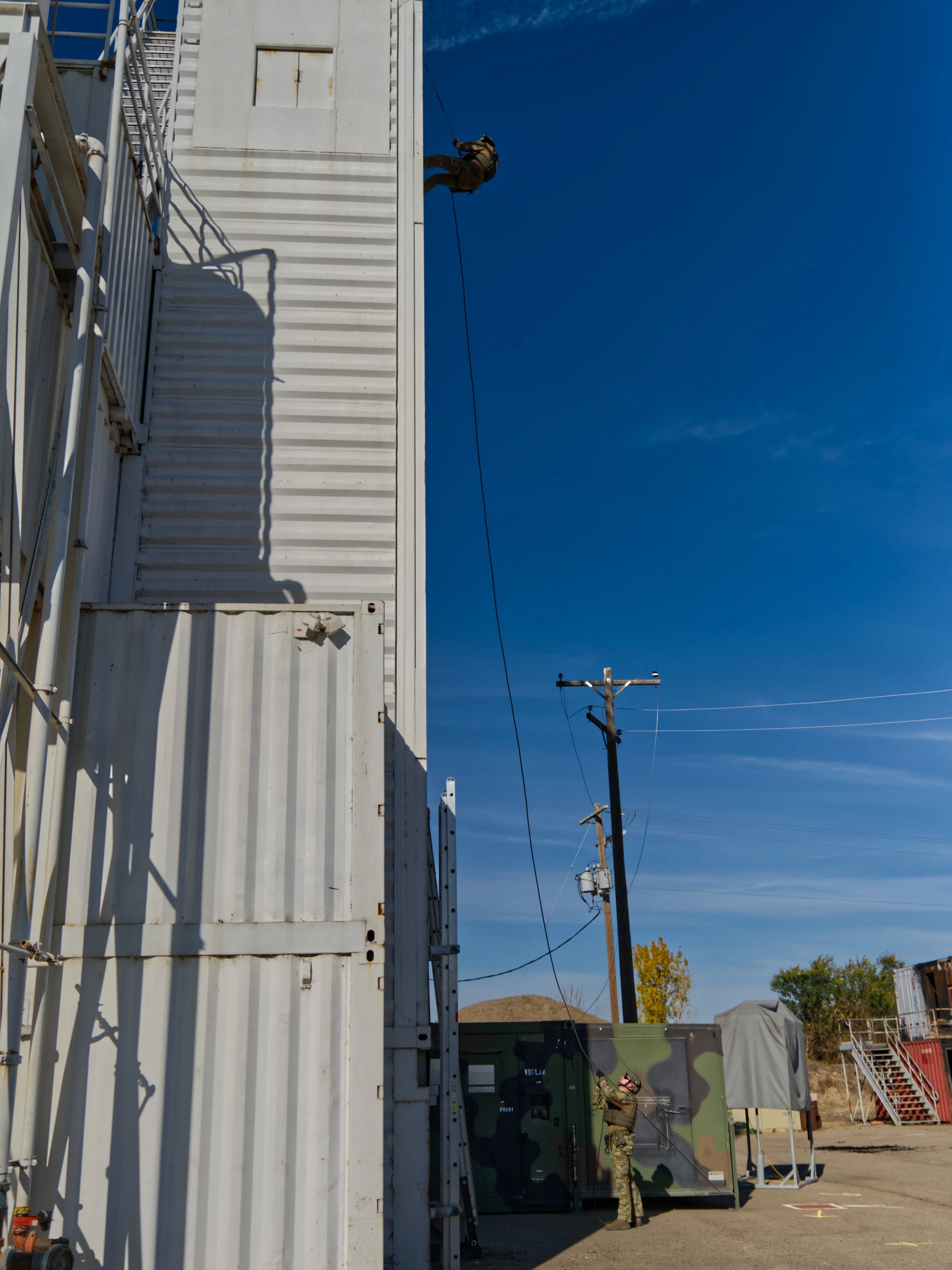 A soldier in camouflage uniform and helmet standing outside near a white container structure, working with equipment, against a bright blue sky with power lines and utility poles in the background.