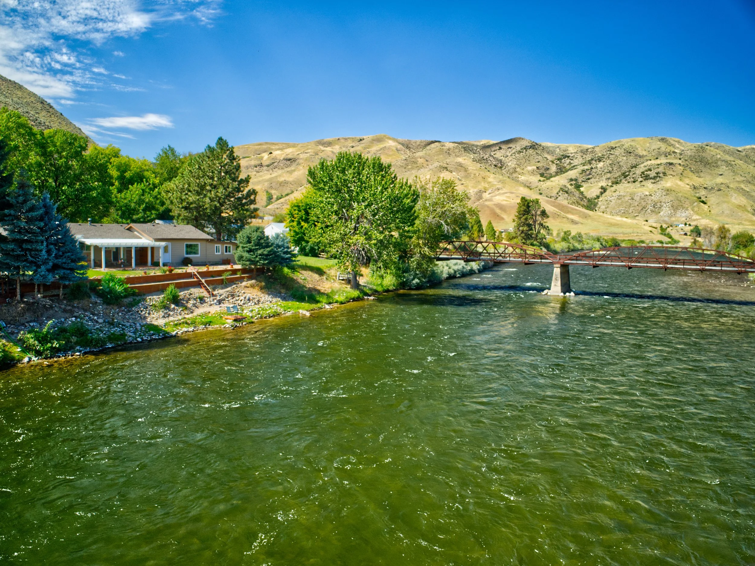 A river flowing through a countryside with green trees, a house, a bridge, and hills under a blue sky with some clouds.