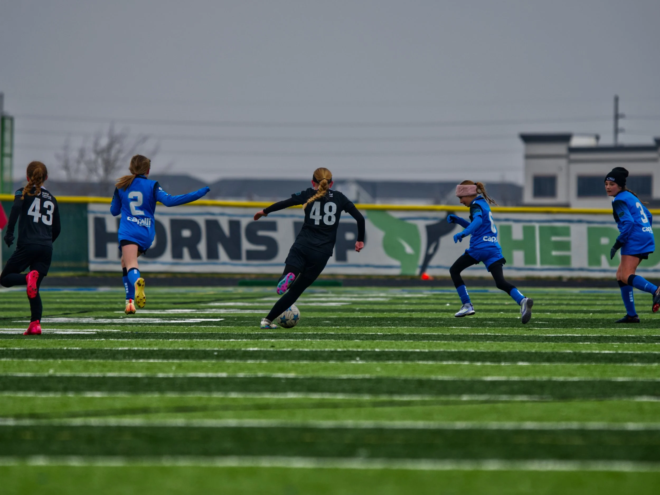 Girls playing soccer on a field during an overcast day, with some wearing black jerseys and others in blue, near the goalpost and advertising boards.