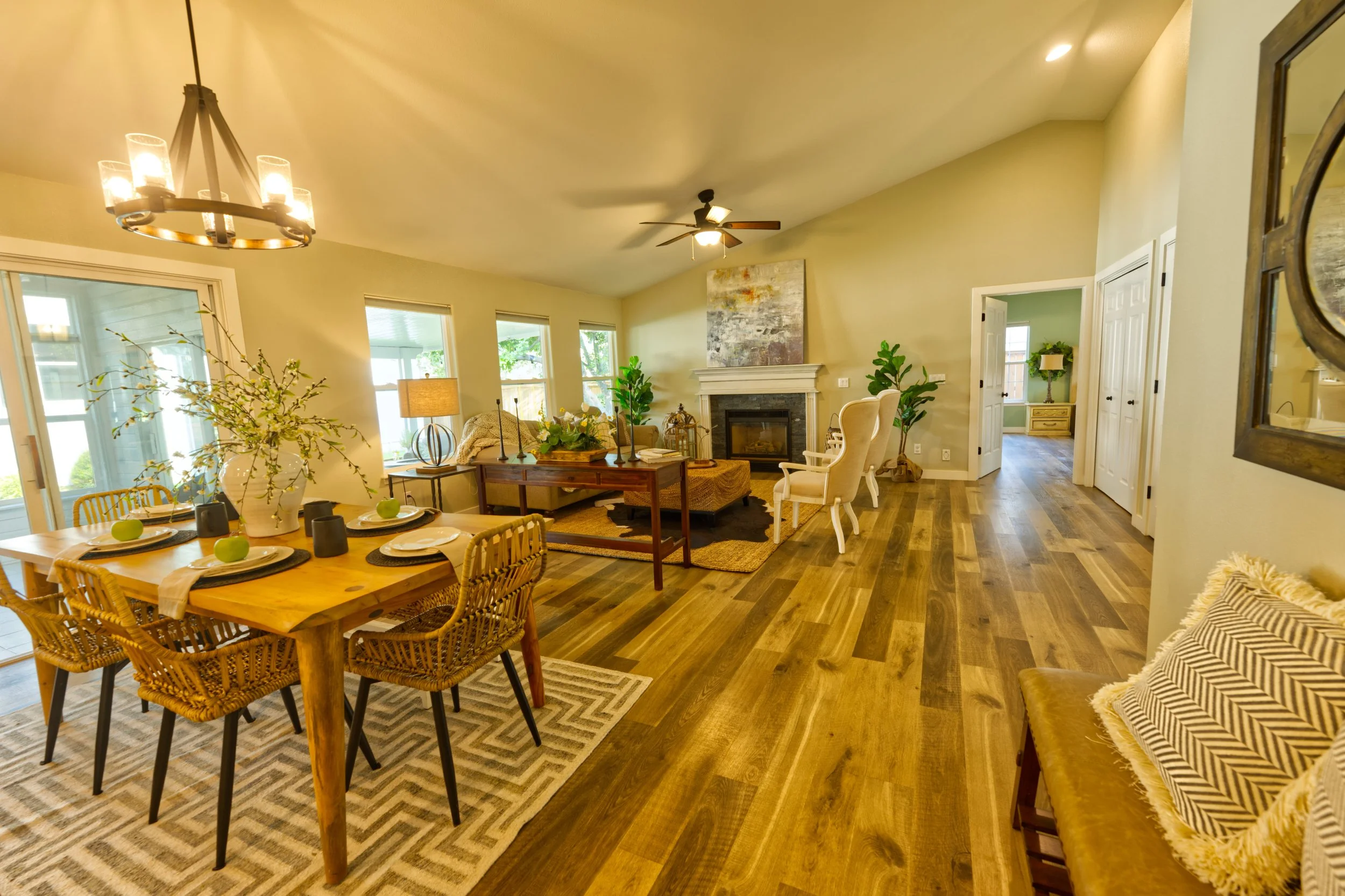 Open-concept living and dining room with wooden flooring, large windows, a front-facing fireplace, ceiling fan, and various seating and decorative items.