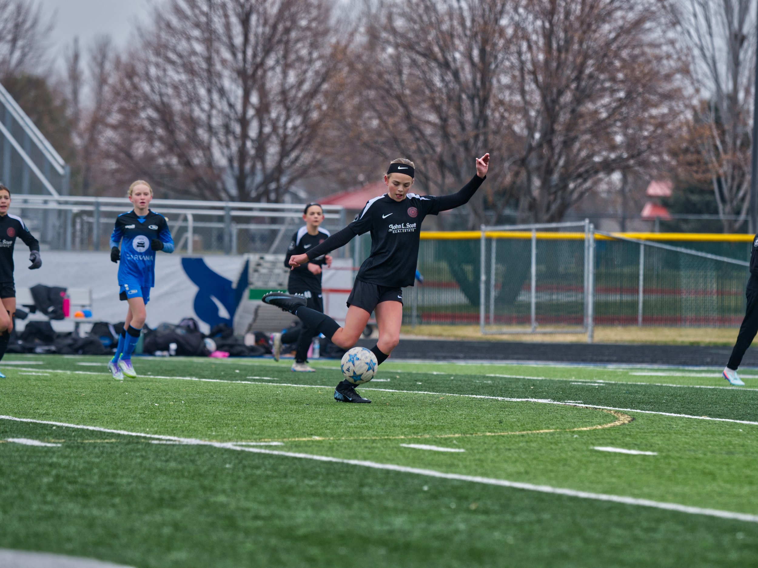 Young girl in a black sports uniform with a Nike headband kicking a soccer ball on a field with trees in the background
