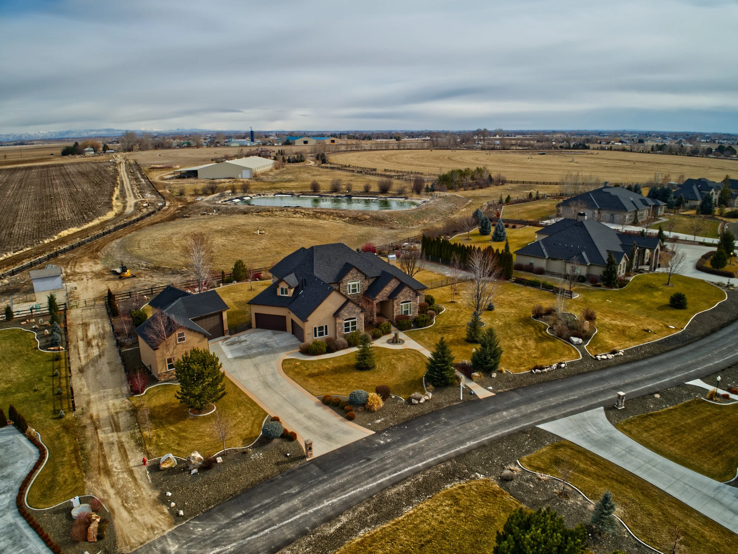 Aerial view of a suburban neighborhood with houses, a pond, and expansive farmland under a cloudy sky.