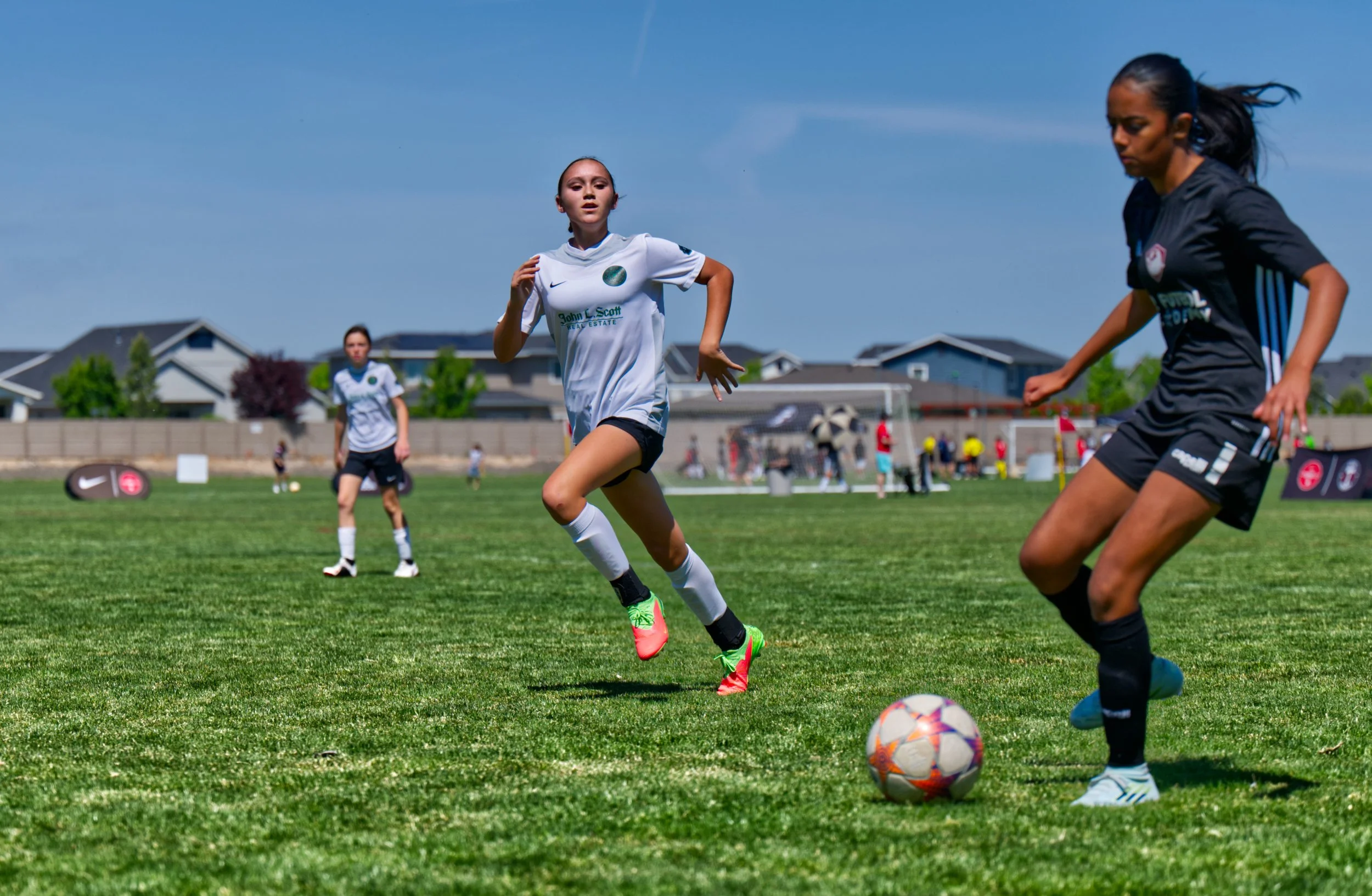 Girls playing soccer on a green field on a sunny day, with houses visible in the background.