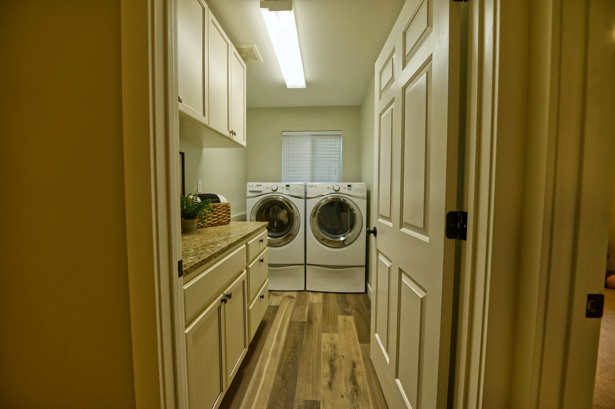 Laundry room with white cabinets, a washer and dryer, a small window, a countertop with a potted plant, and a wicker basket, viewed through a doorway.