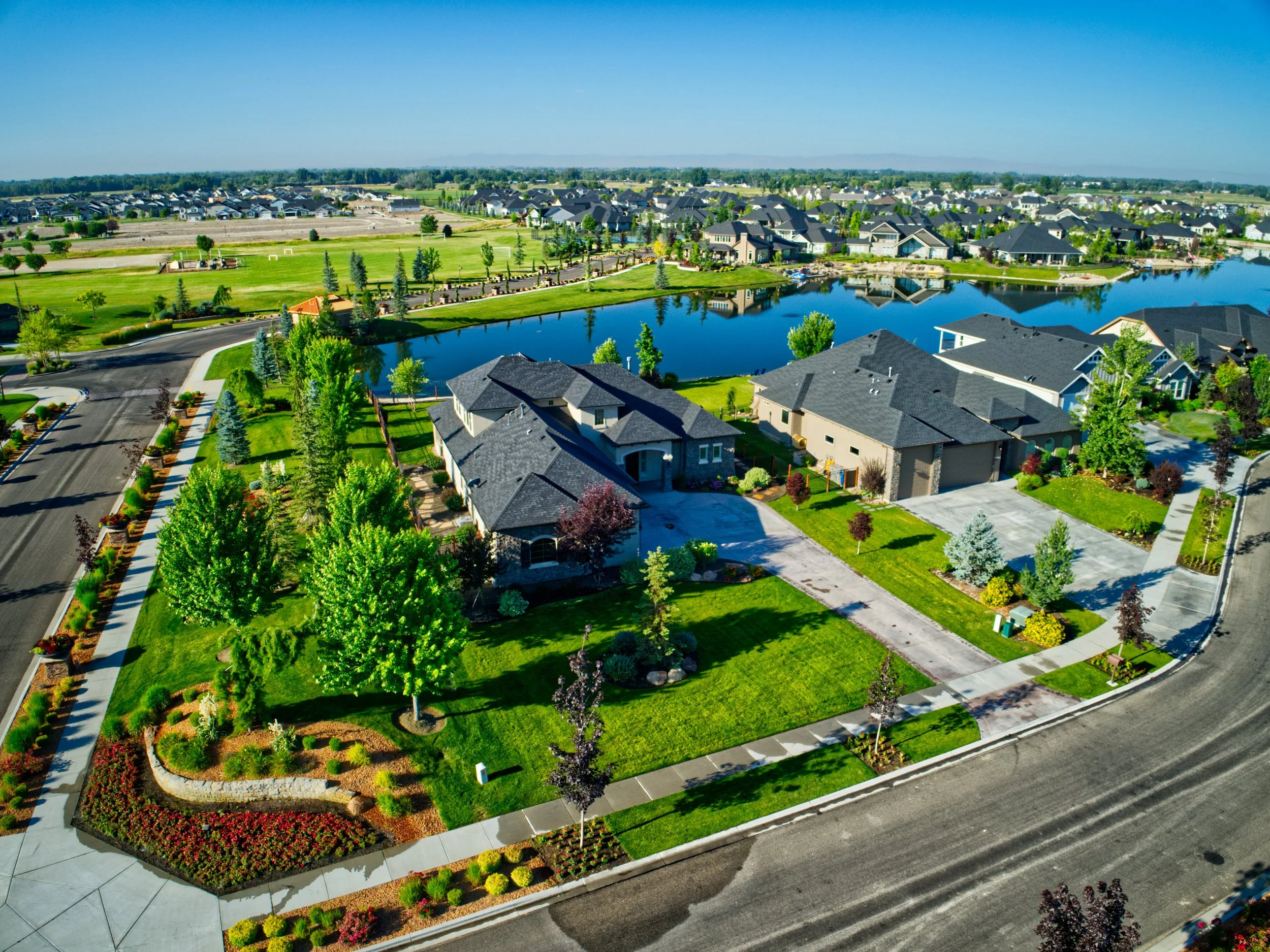 Aerial view of a suburban neighborhood with a lake, houses, green lawns, roads, and trees under a clear blue sky.