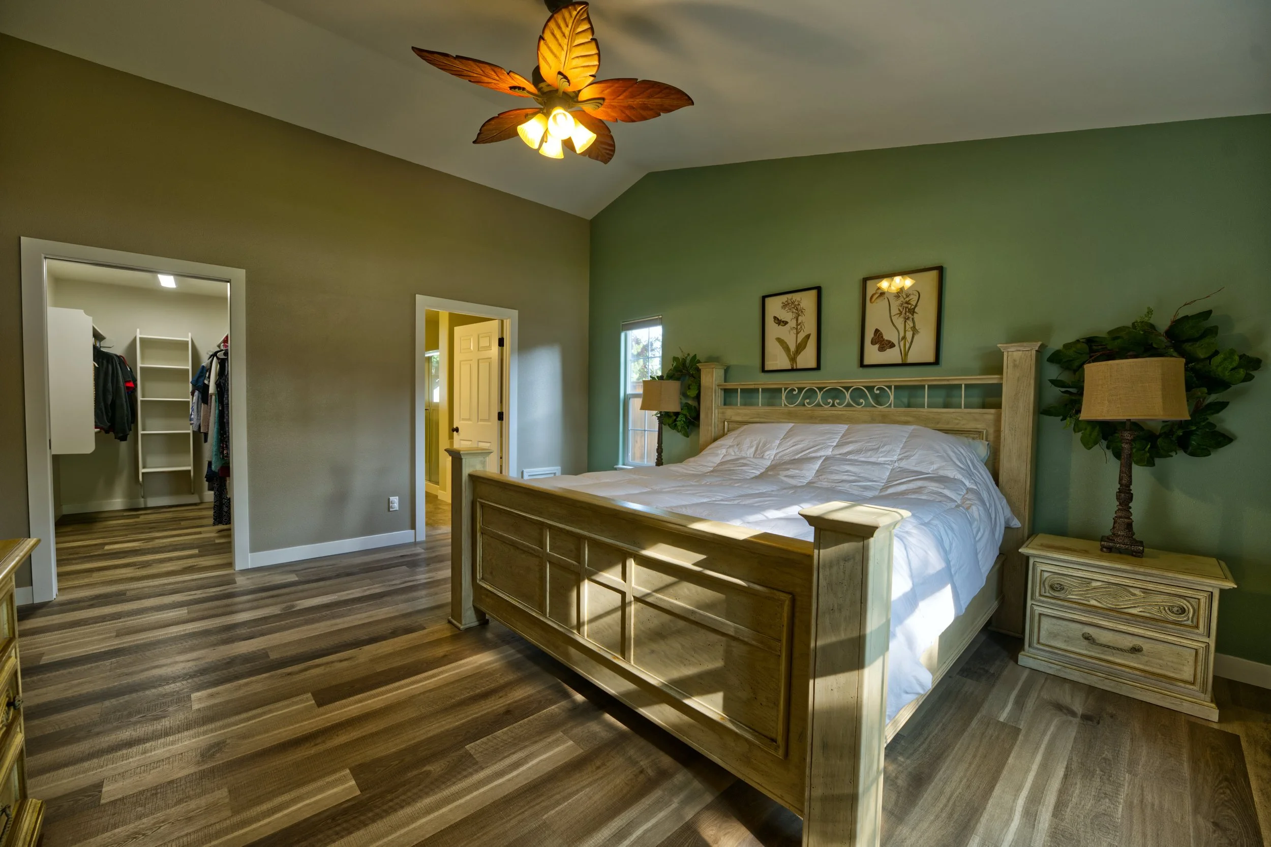 A bedroom with a wooden bed frame, nightstand, and a lamp on each side, with two framed botanical art prints on the wall, and a ceiling fan with leaf-shaped blades.