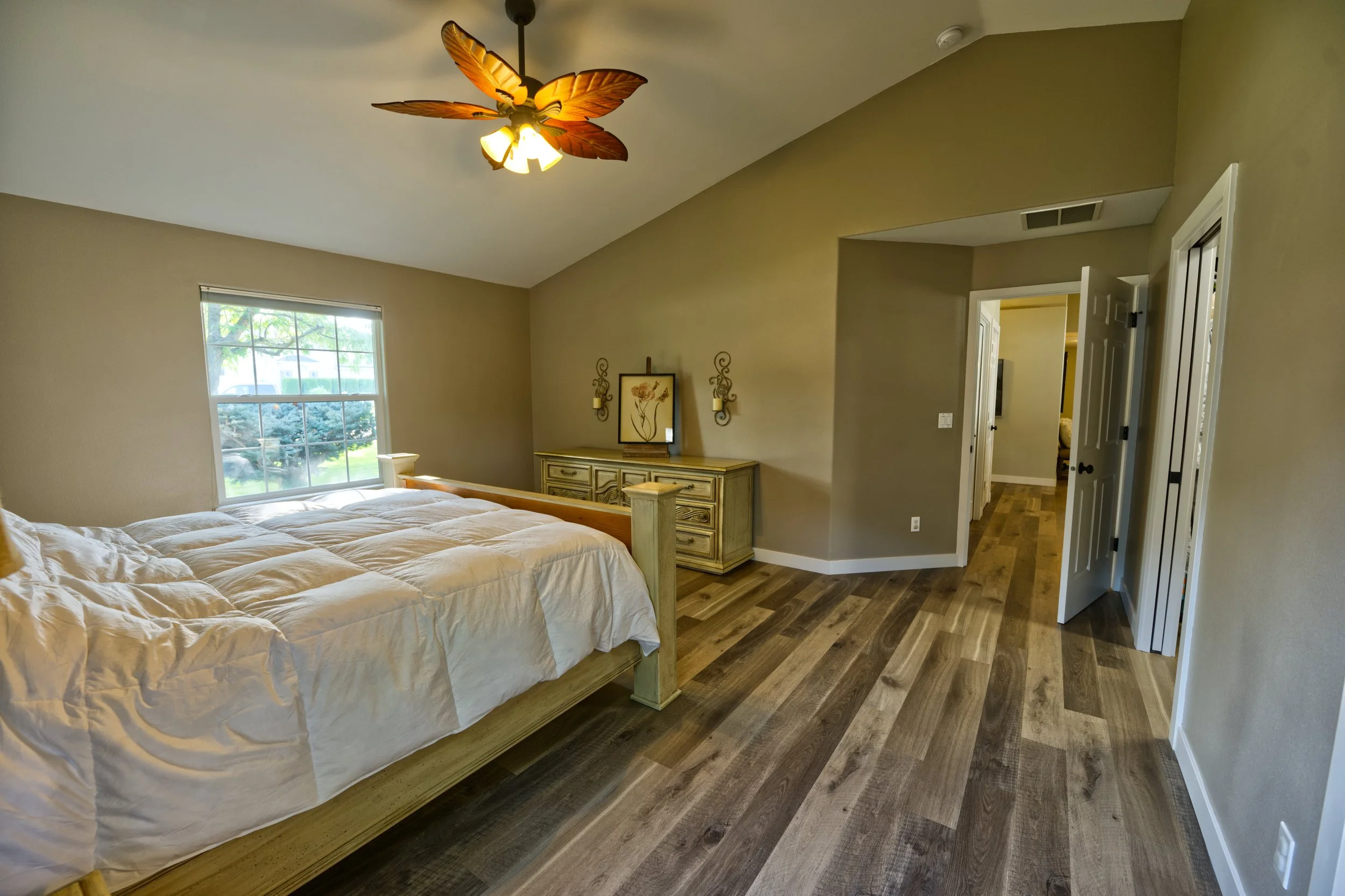 A bedroom with a large bed with white bedding, a window showing a green yard outside, a ceiling fan with butterfly-shaped blades, and a wooden dresser with a framed art piece and wall-mounted candle holders.