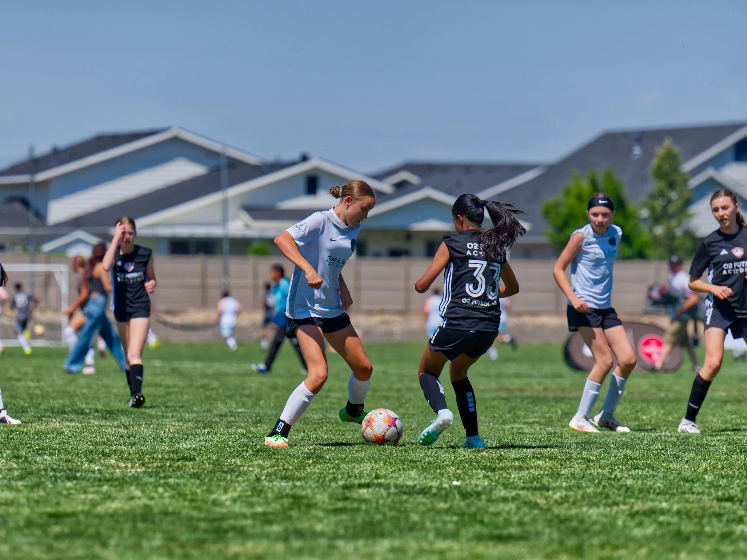 Young girls playing soccer on a grass field on a sunny day, with residential houses in the background.