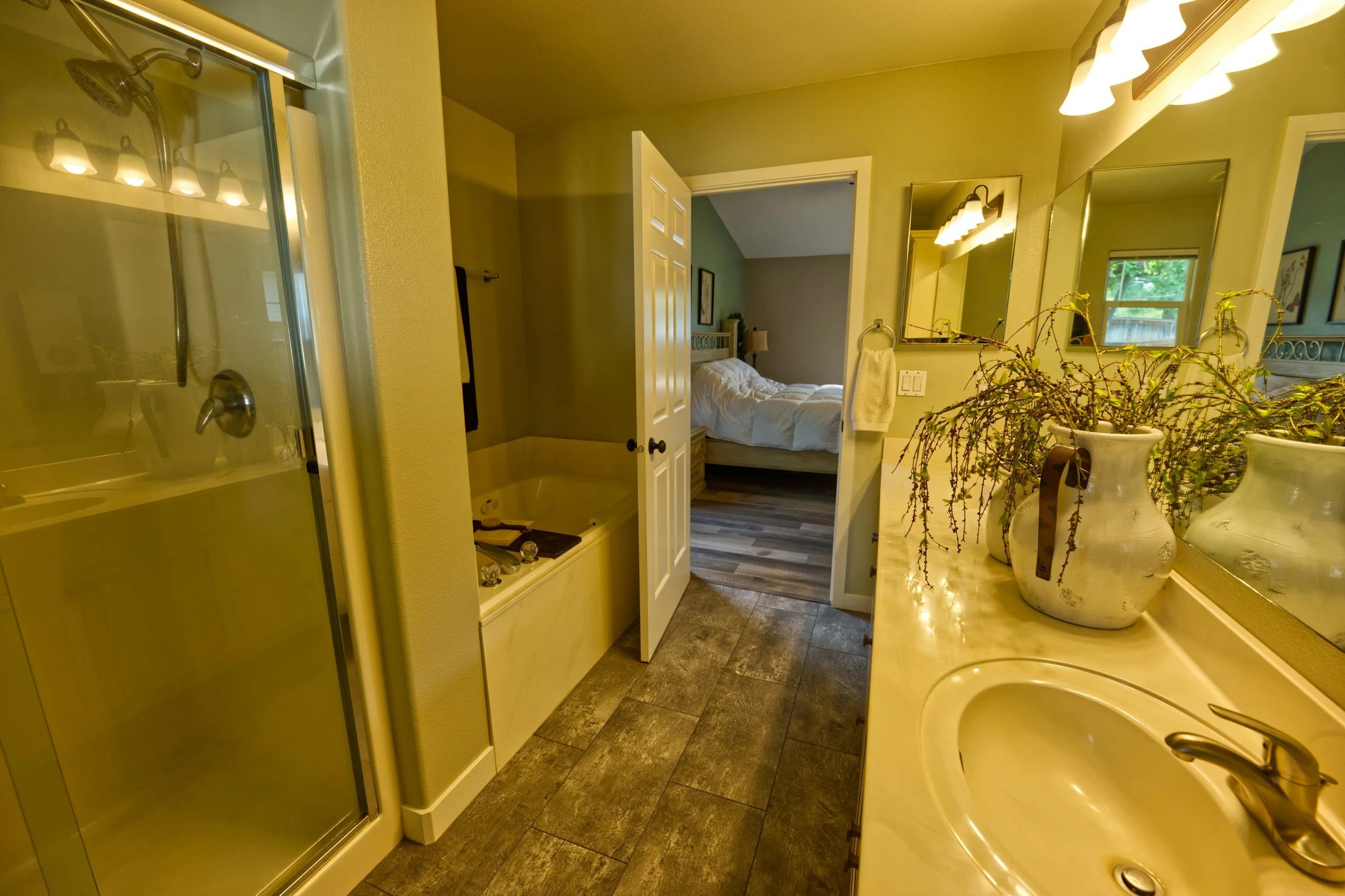 Bathroom with a shower stall on the left, a bathtub below the middle, and a double sink with a large mirror on the right. Tucked into an open doorway is a bedroom with a bed, nightstand, and window.