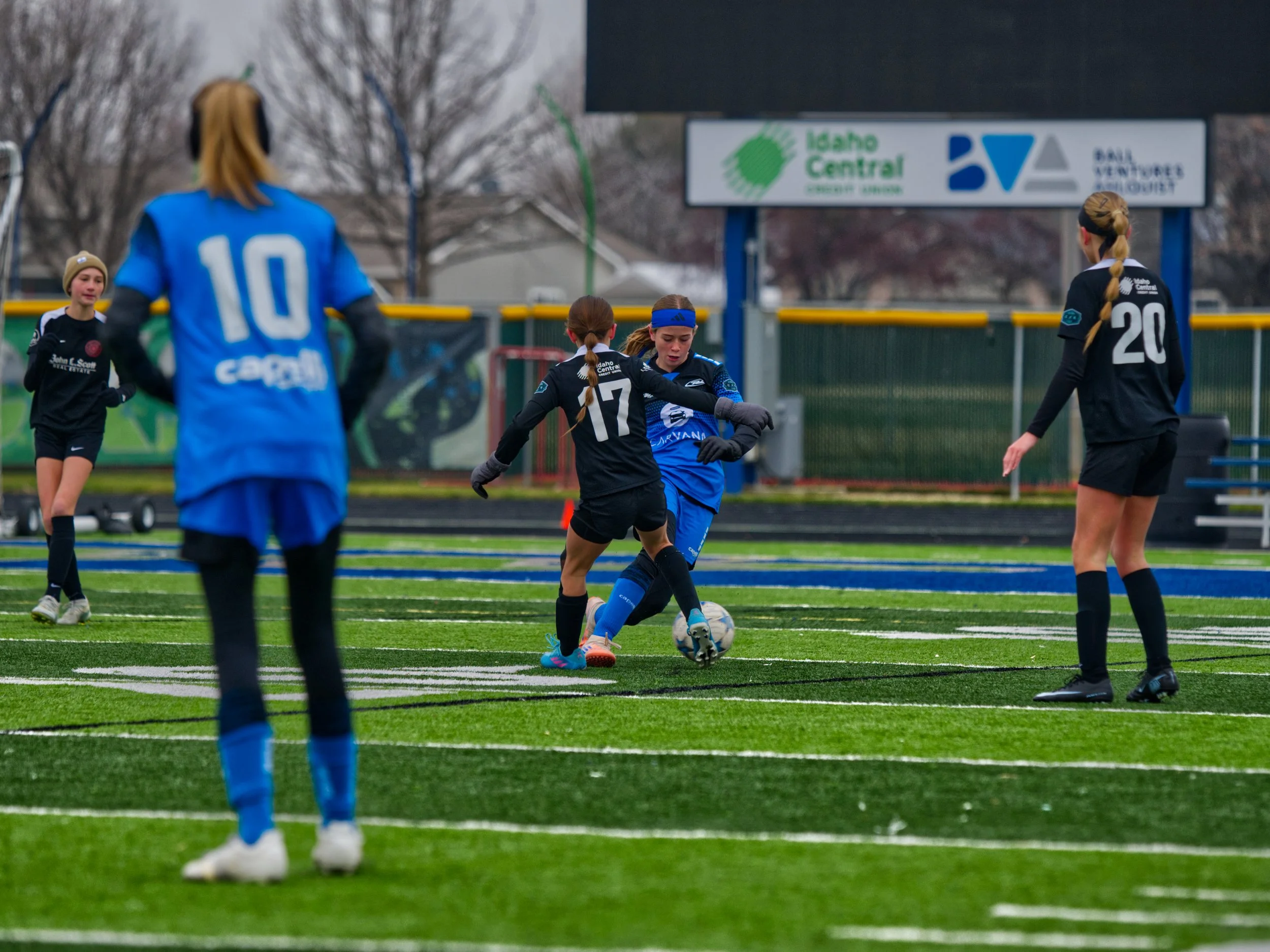 Girls playing soccer on an outdoor field, some in black uniforms and one in a blue shirt with the number 10, some wearing headbands and gloves, with trees and a sign in the background.