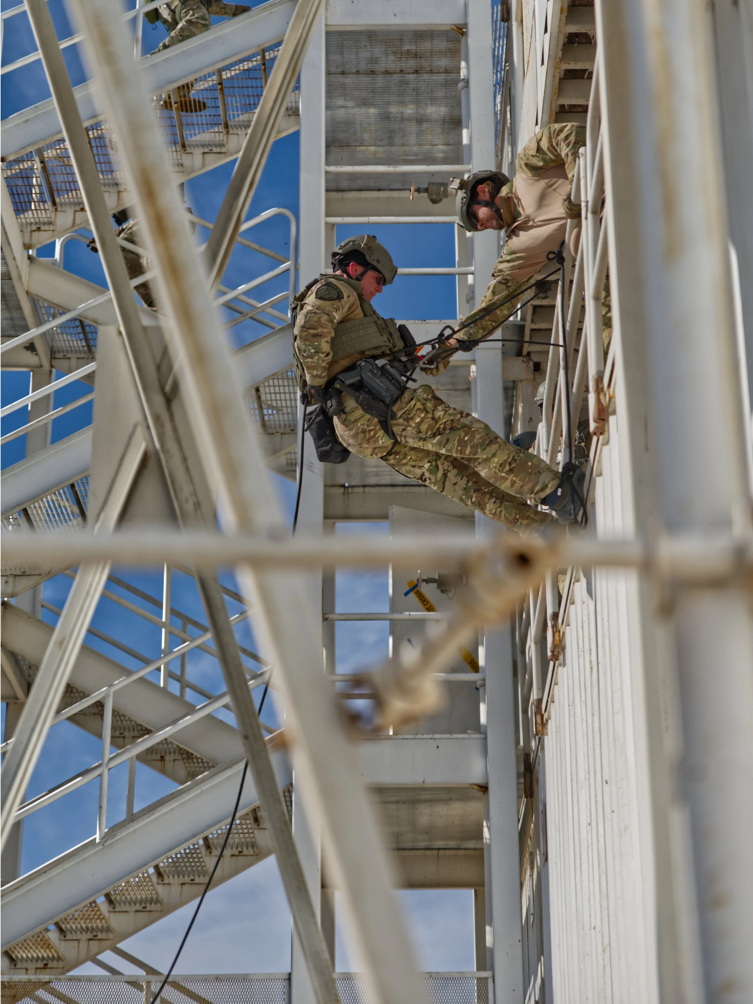 Two soldiers wearing camouflage uniforms and helmets are working on a metal staircase structure outdoors against a blue sky. One soldier is climbing while the other is assisting by holding a rope or cable.