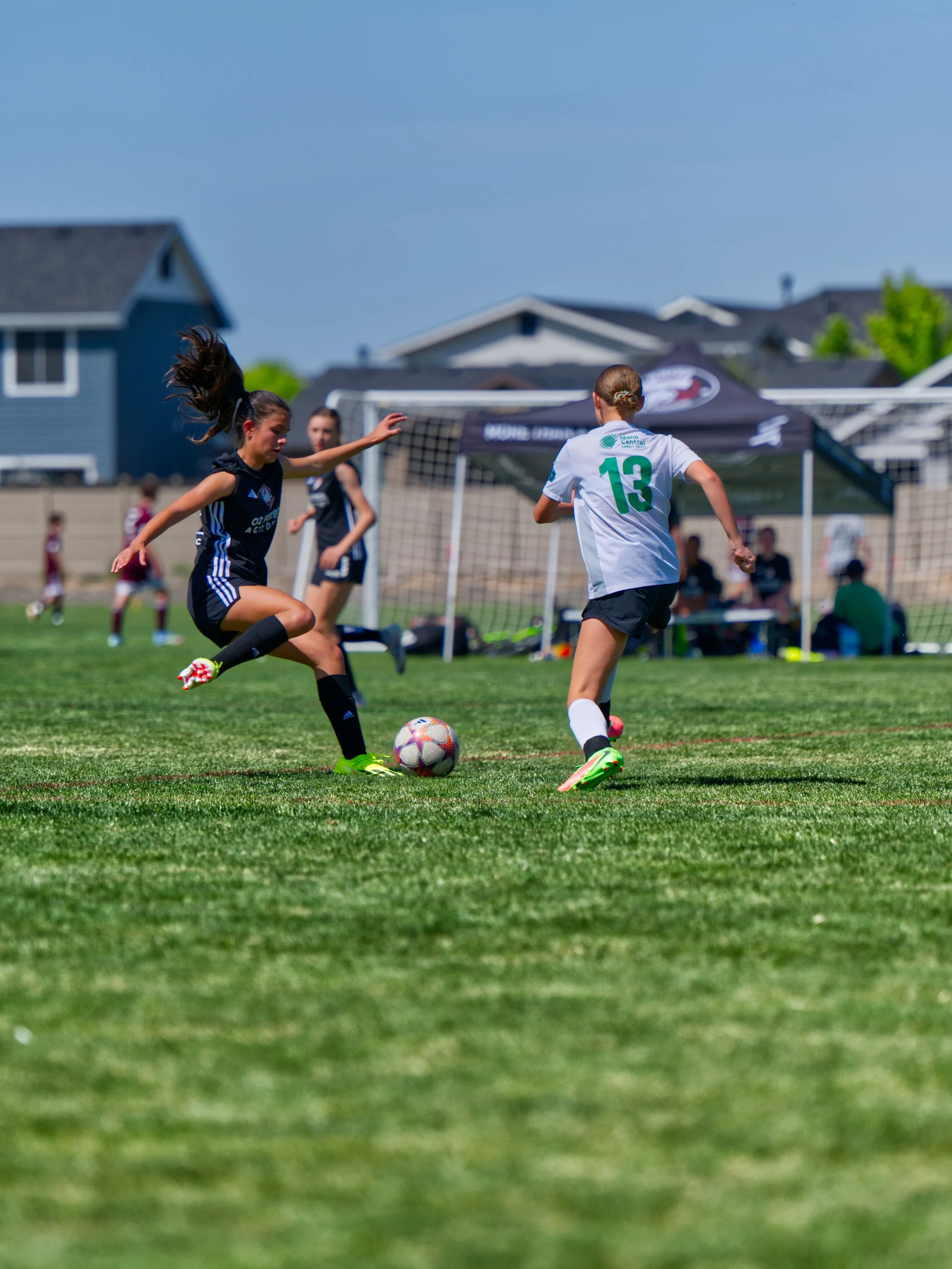 Two female soccer players are competing for the ball on a grassy field during a daytime match. One player is wearing a black uniform and the other a white uniform. In the background, there is a goal, a canopy, and several spectators.
