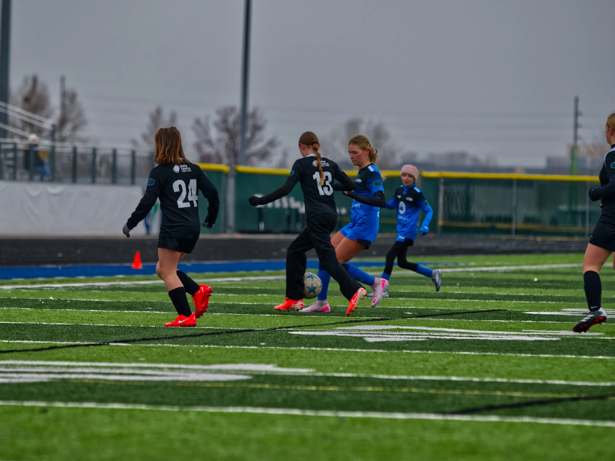 A group of young female soccer players on an outdoor field, dressed in black and blue uniforms, competing for the ball under a cloudy sky.