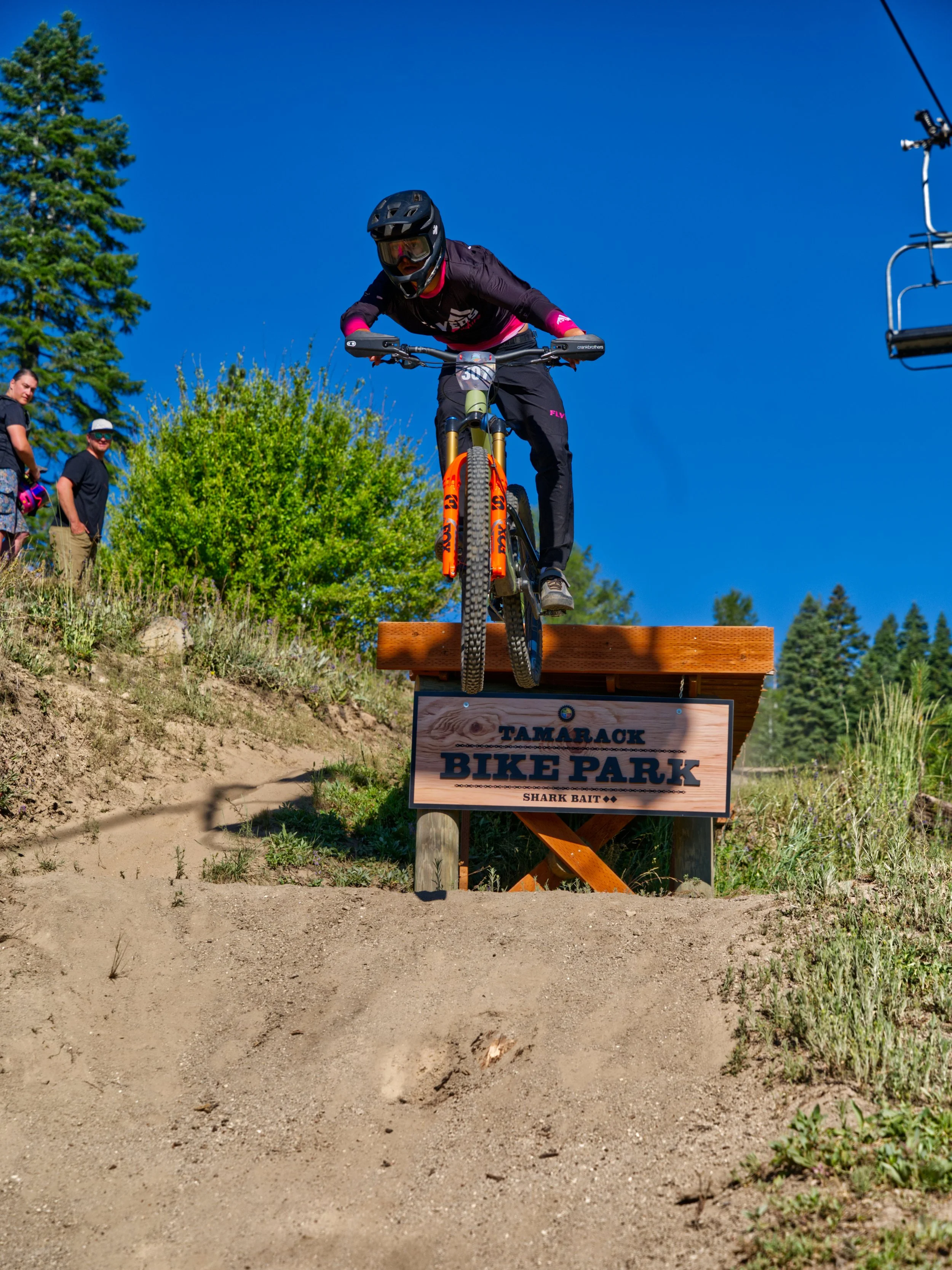 A person riding a mountain bike off a wooden ramp at Tamarack Bike Park on a sunny day, with two people watching nearby and trees in the background.