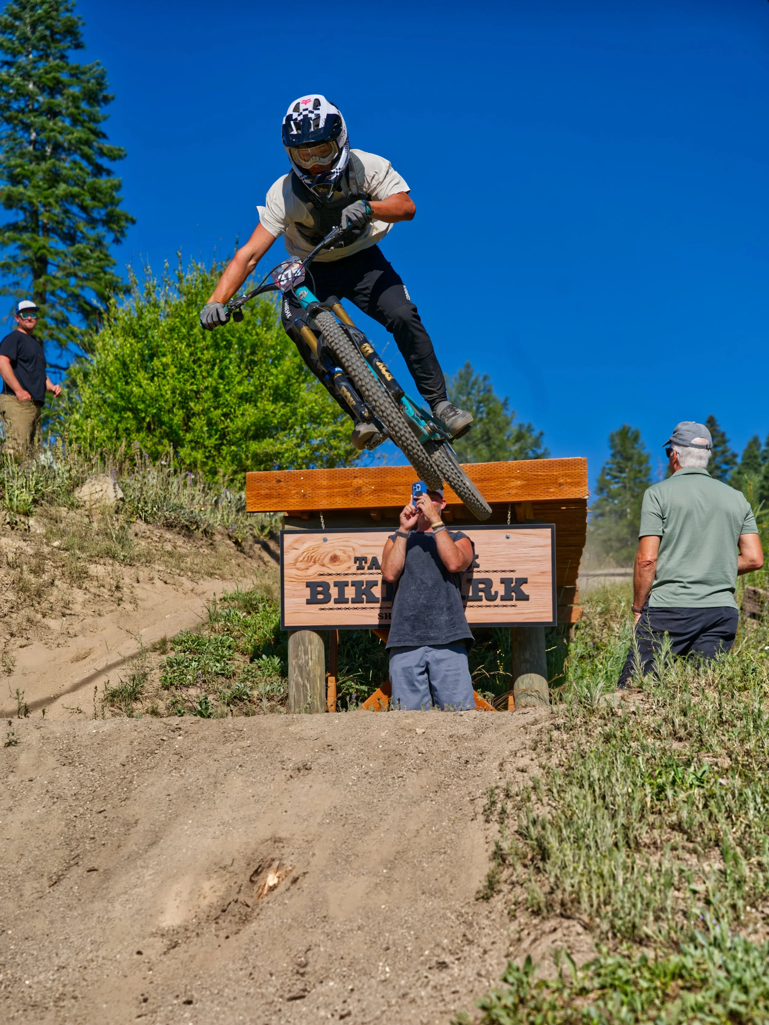 A mountain biker in a white shirt and black pants riding down a dirt slope at an outdoor trail park on a sunny day. A man is taking a photo of the rider from below, and others are watching from the side.