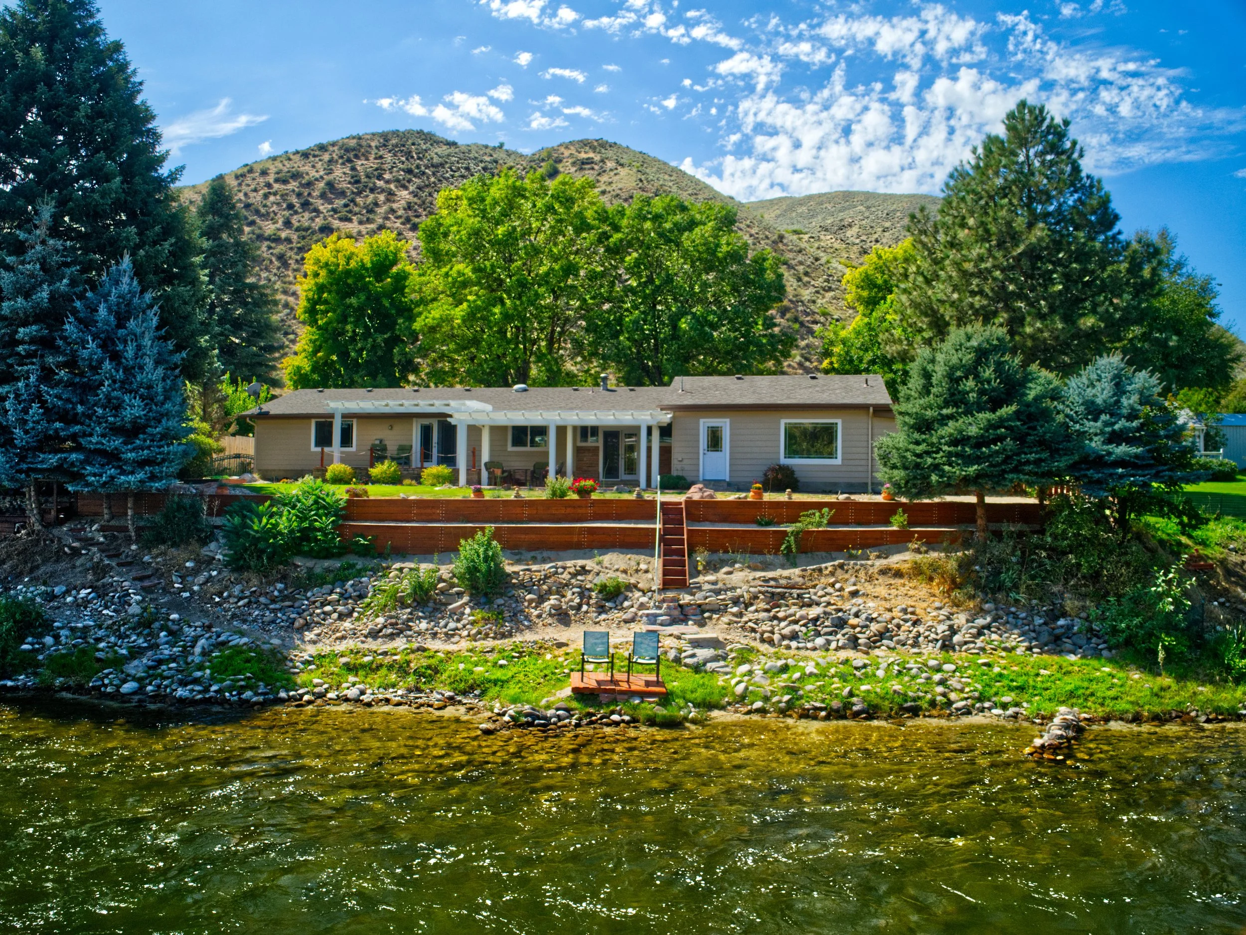 A house near a river with a mountain and blue sky in the background, surrounded by trees and a landscaped yard.