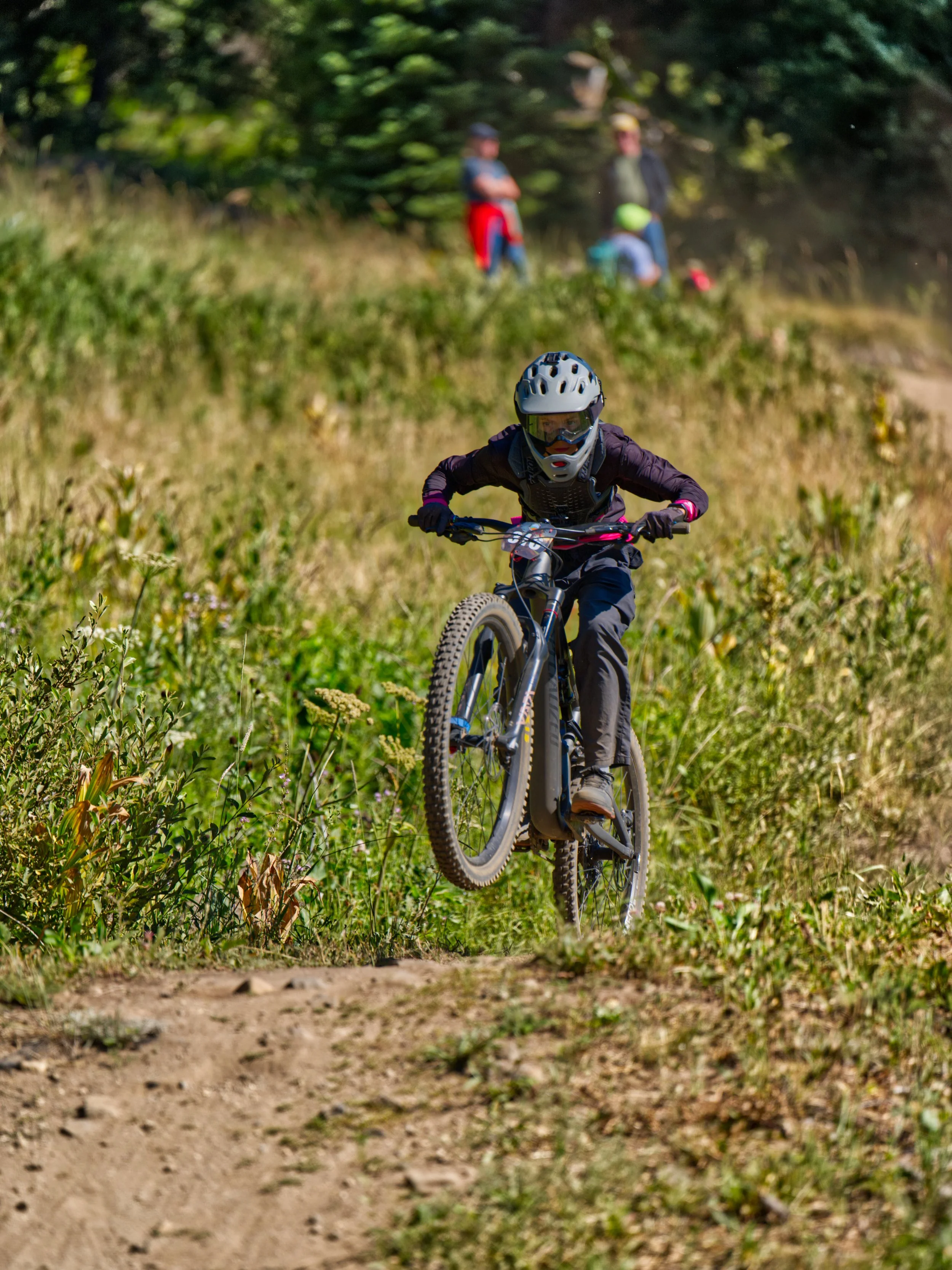 A person wearing a helmet and protective gear riding a mountain bike on a dirt trail through a grassy field.