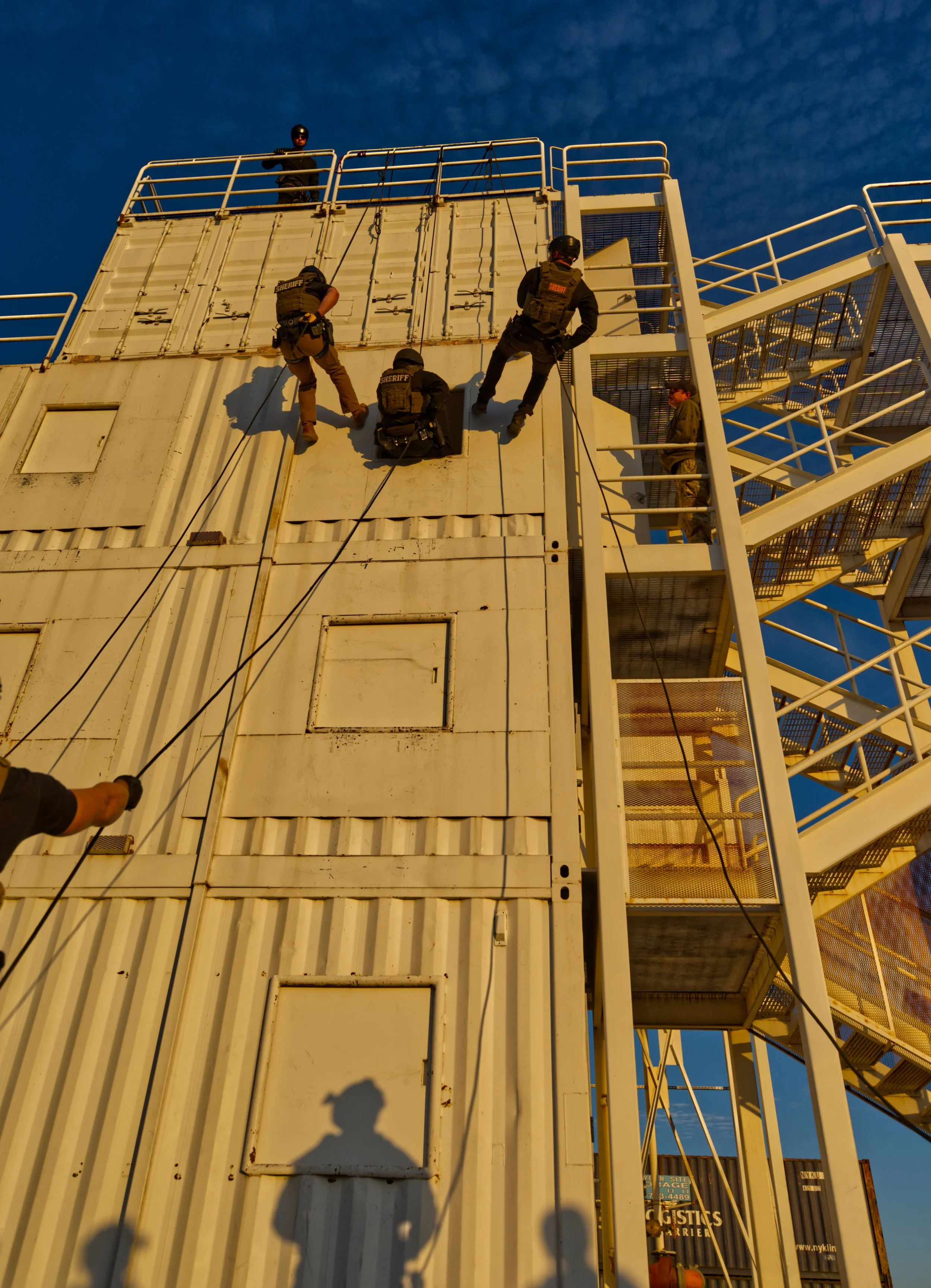 Four sheriff deputies rappelling down a large white shipping container with a staircase on the right and a blue sky background.