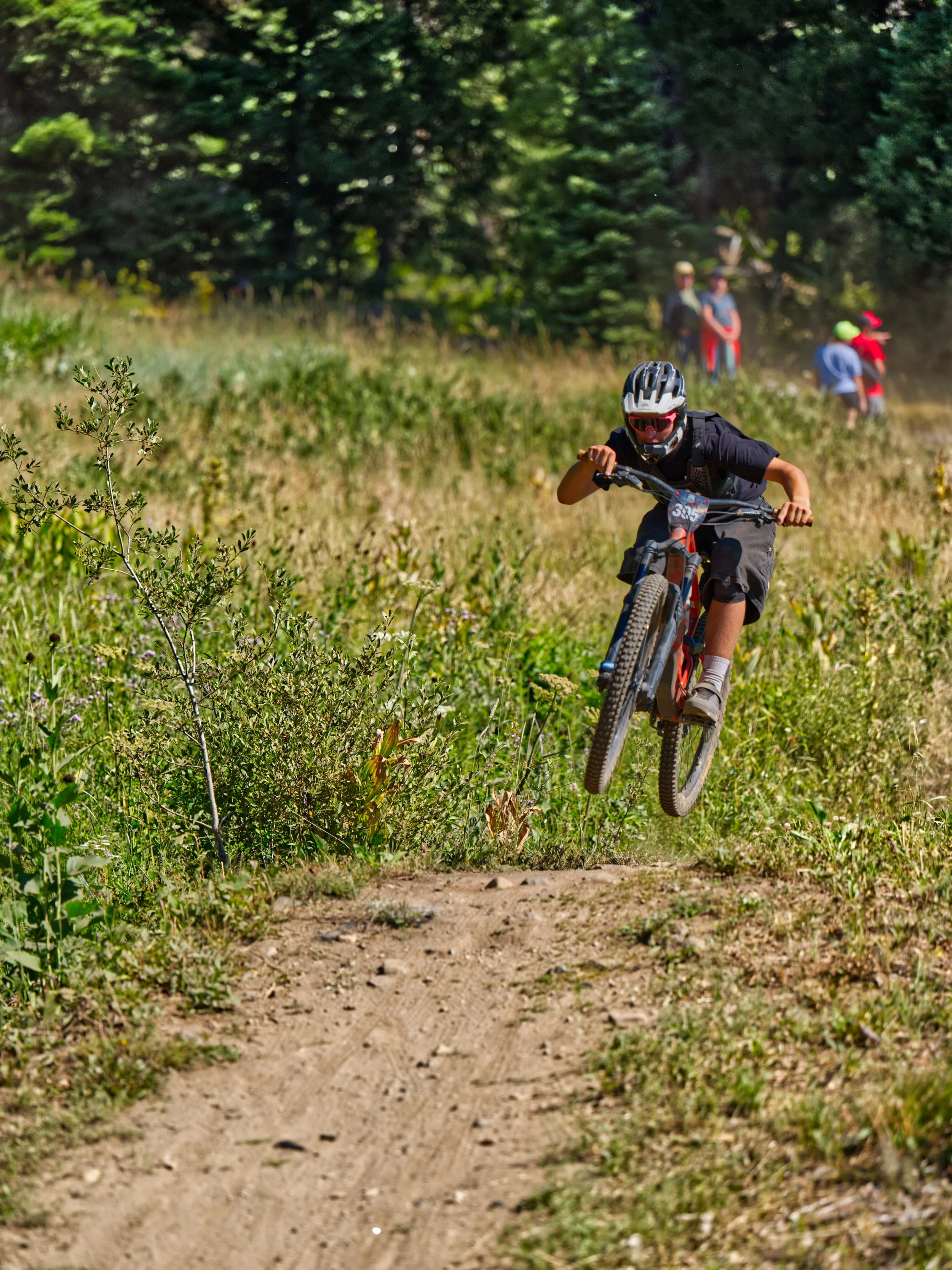 A young boy riding a mountain bike on a dirt trail in a forest. He is wearing a helmet, goggles, and black clothing. In the background, there are several people standing on the trail, some wearing helmets and casual clothing, near tall green trees an