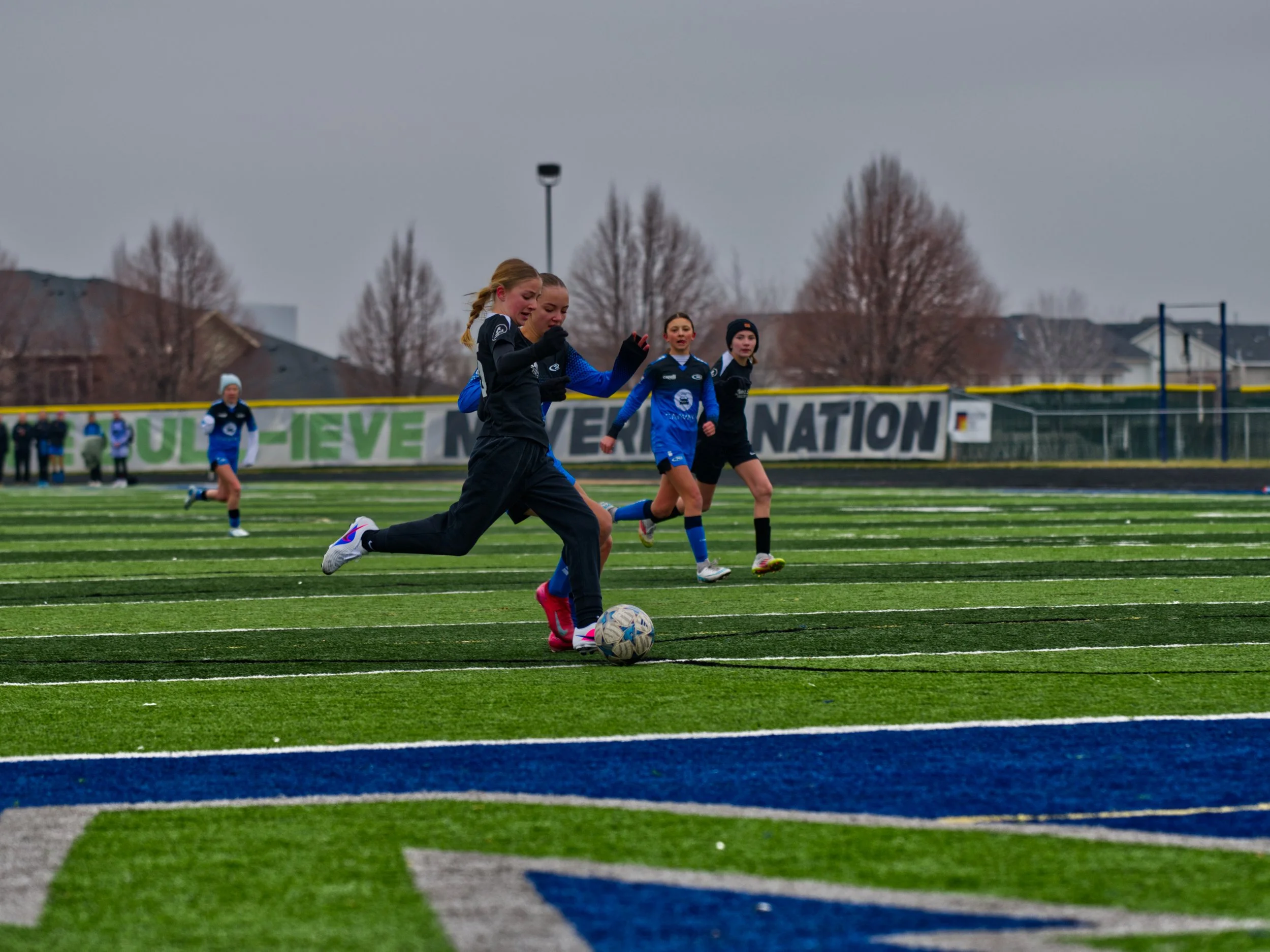 Girls playing soccer on a field with yard lines, wearing sports uniforms, during cloudy weather.
