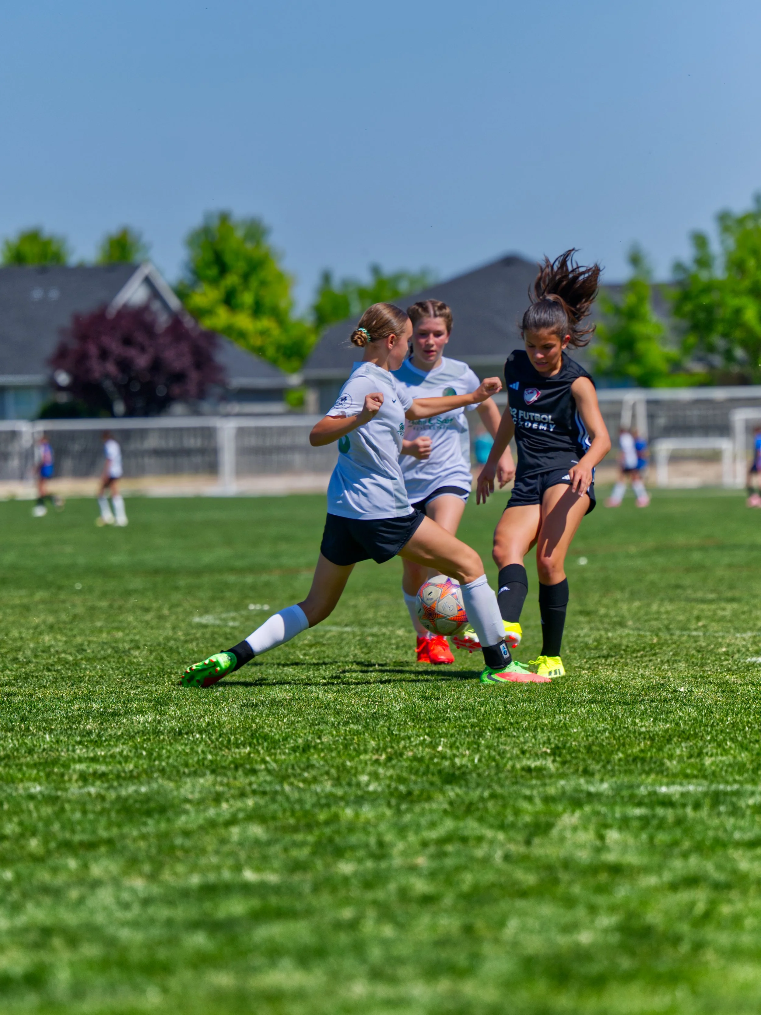 Three young girls playing soccer on a green field under a clear blue sky, with houses and trees in the background.