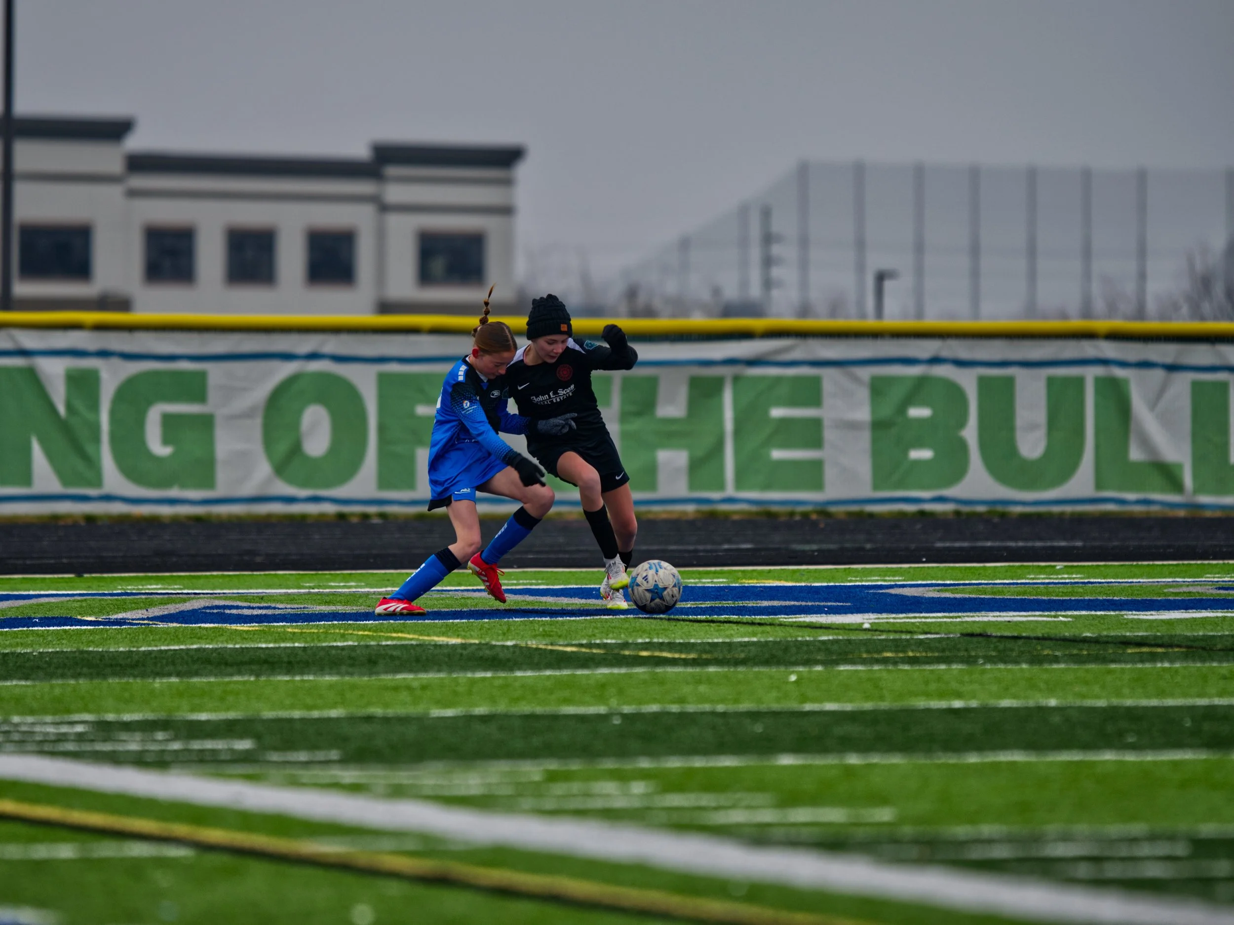 Two young girls playing soccer on a field, competing for the ball, with a sports fence and buildings in the background on a cloudy day.