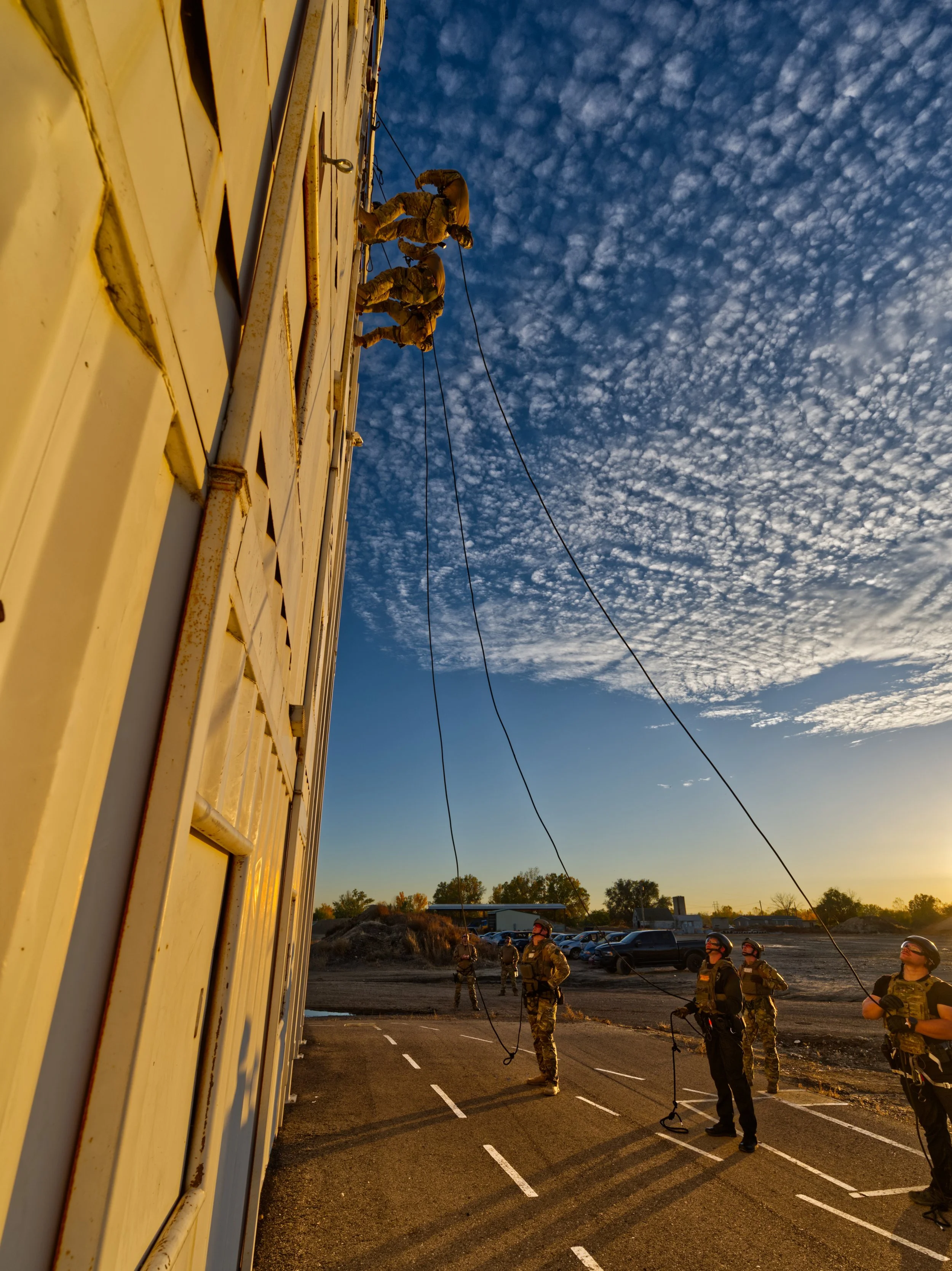 Image of soldiers standing near a large yellow structure, possibly on a military base, during sunset or early morning with a cloudy sky and a parking lot in the background.