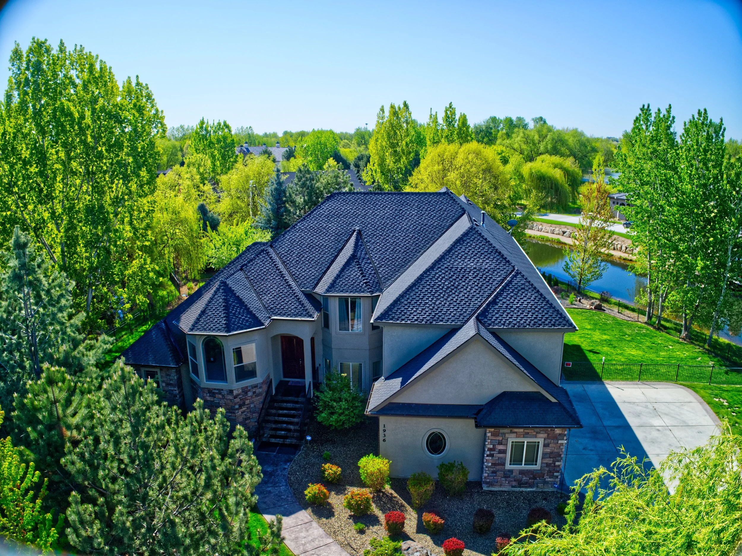 A large house with gray roof shingles and brick accents, surrounded by tall green trees and colorful shrubs, with a pond and a driveway in a lush landscape.