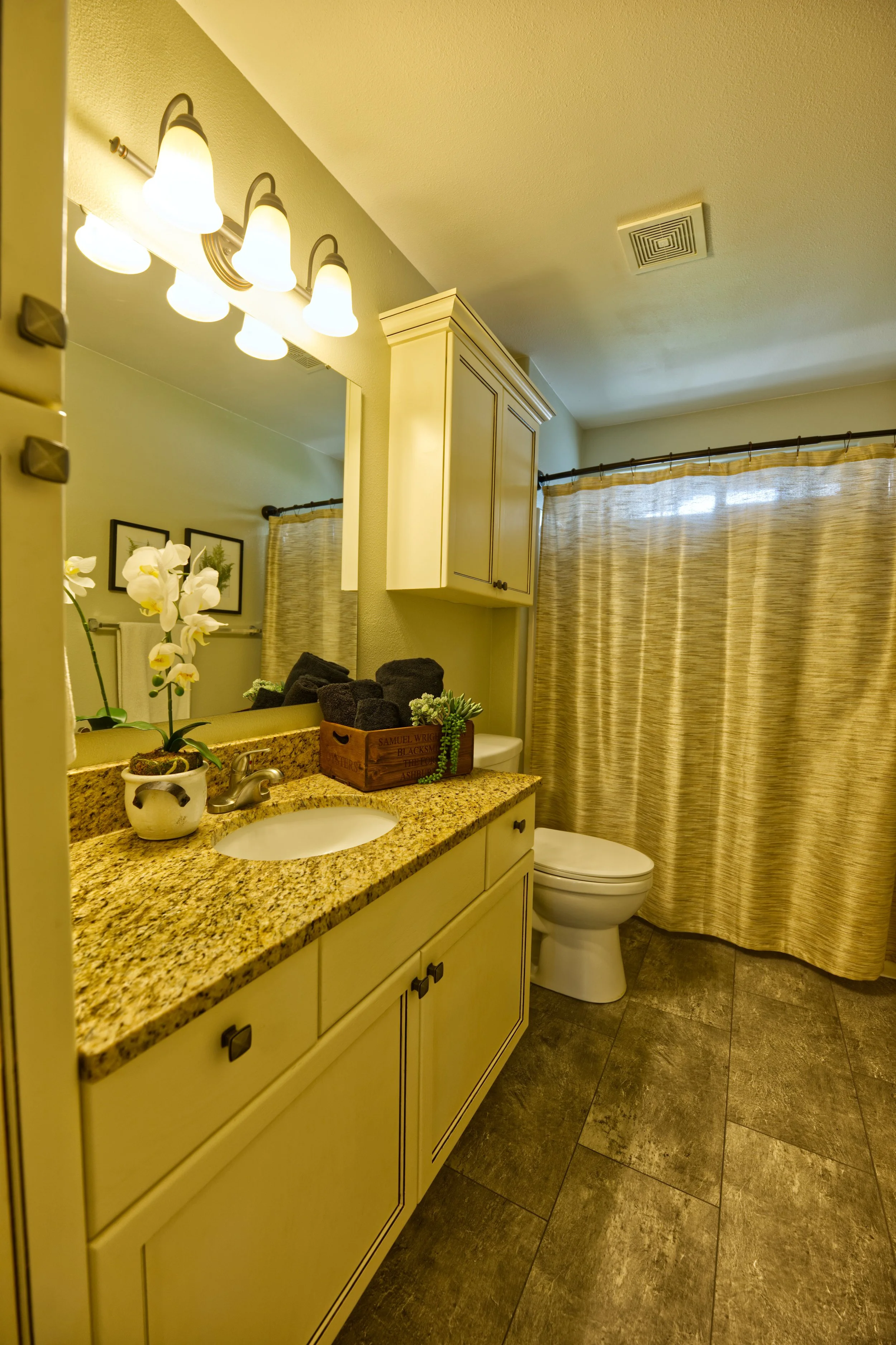 A bathroom with a granite countertop, a white sink, a mirror, floral decor, a white cabinet, a toilet, window with a beige striped curtain, and a ceiling vent.