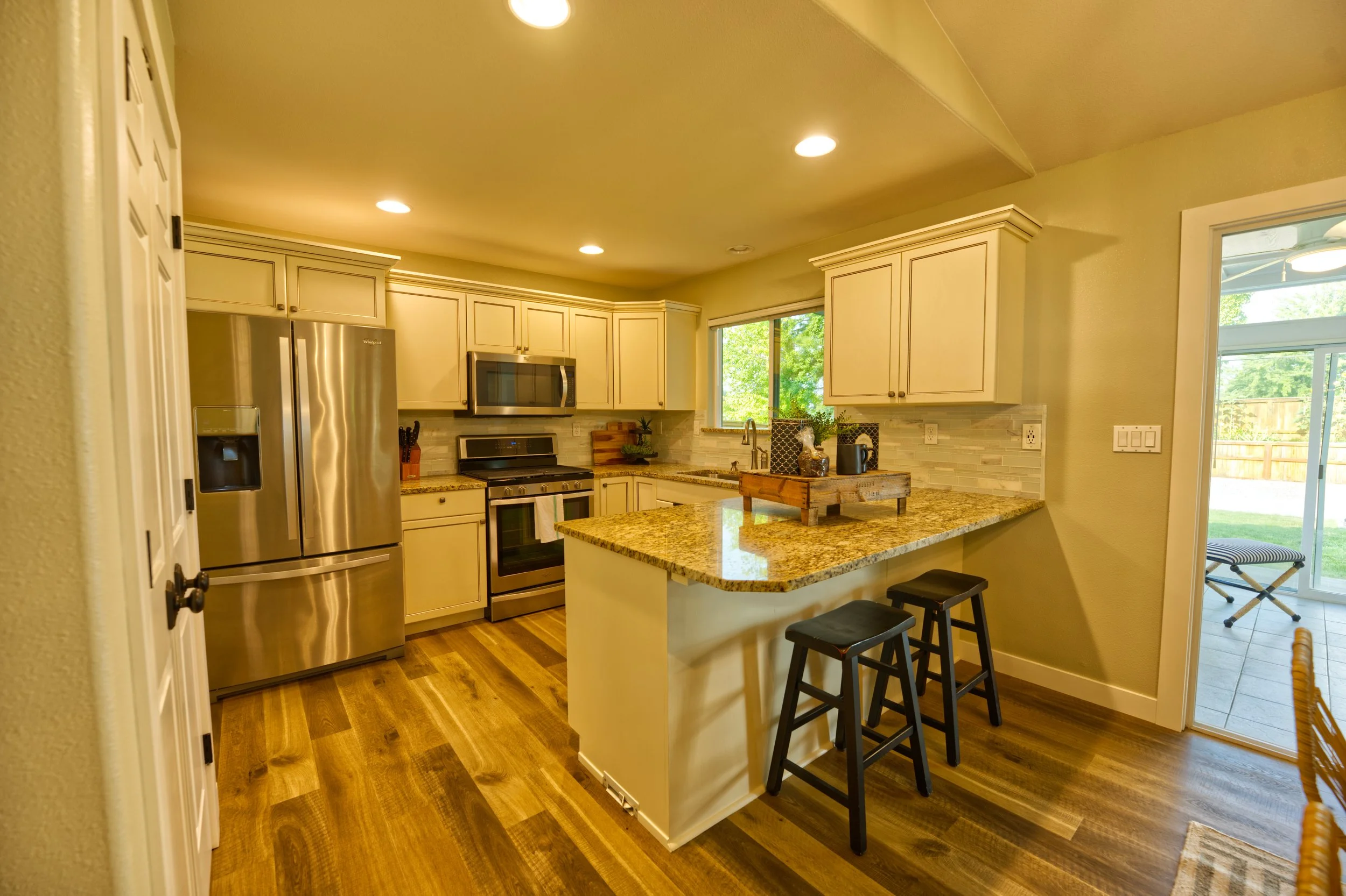 A modern kitchen with beige cabinets, a stainless steel refrigerator, a microwave, and a granite countertop island with three black stools. There is a window with a view of greenery and a sliding glass door leading outside, with natural light filling