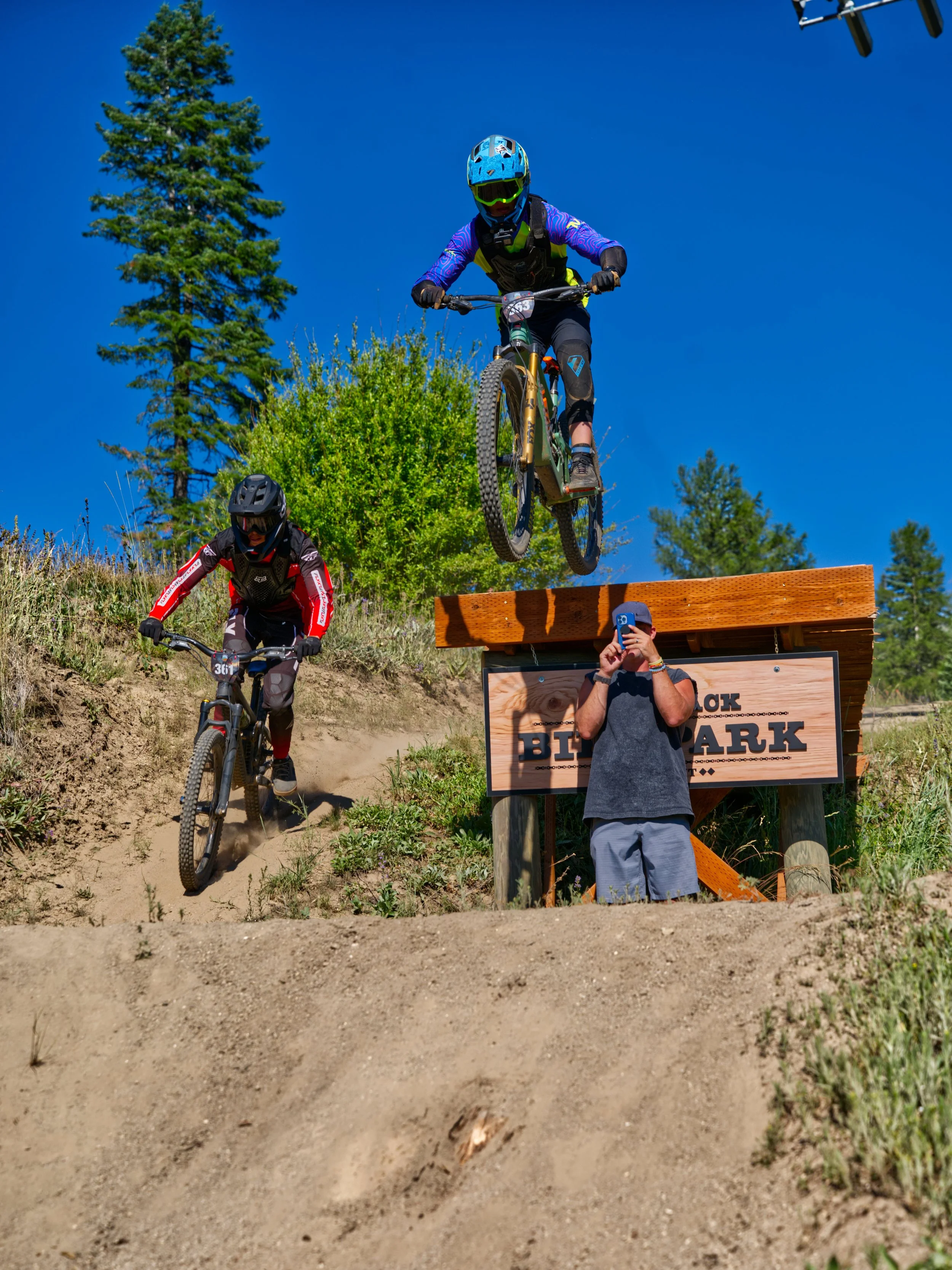 A mountain biker is mid-air jumping off a wooden ramp at a dirt trail, with two other cyclists nearby and a person taking a photo or video underneath the ramp. The scene is outdoors with trees and a clear blue sky.