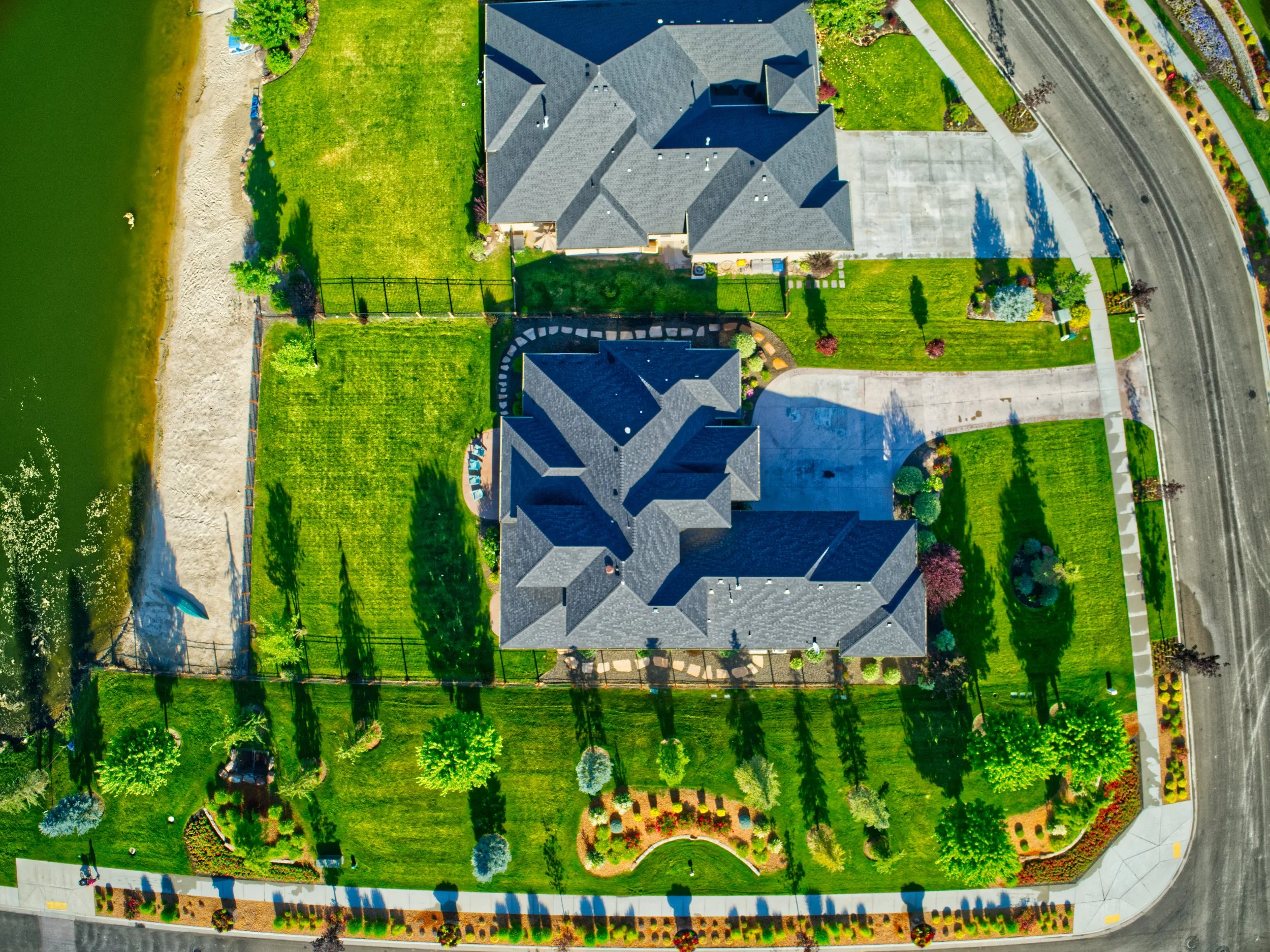 An aerial view of a residential property with a large house featuring a gray roof, a well-maintained lawn, a driveway, and landscaped gardens along a curved street.