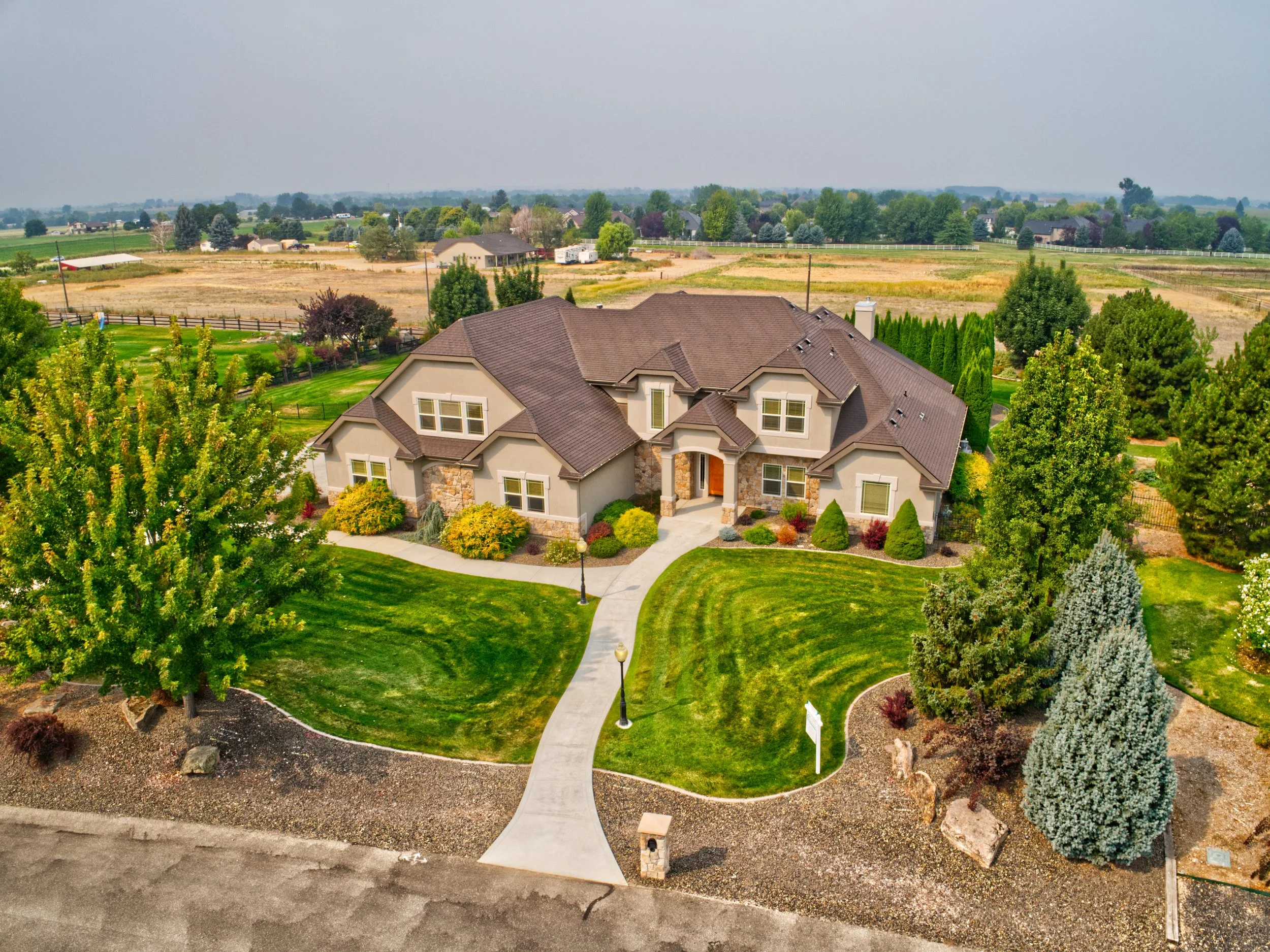A large house with a tan and brown roof, surrounded by a well-maintained green lawn and trees, leading to a sidewalk and a street with a mailbox, set in a suburban neighborhood with open fields and additional houses in the background.
