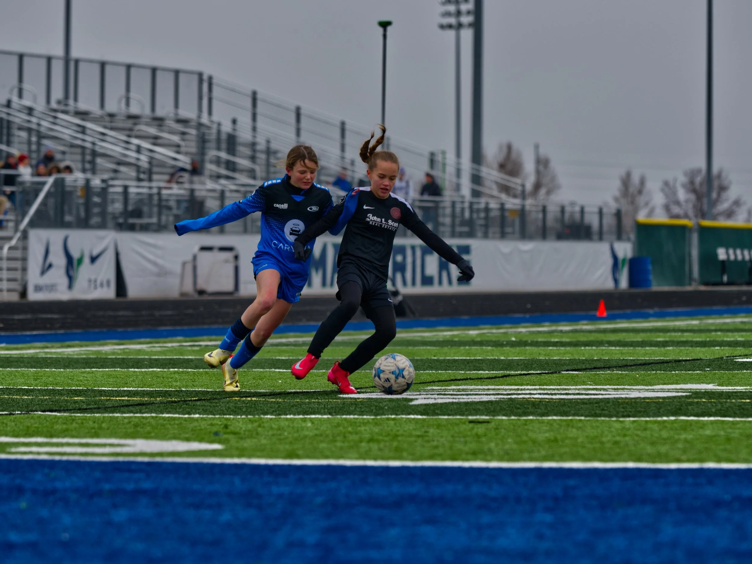 Two young girls are competing in a soccer game on a turf field, chasing after a soccer ball, with stands and light poles in the background on a cloudy day.