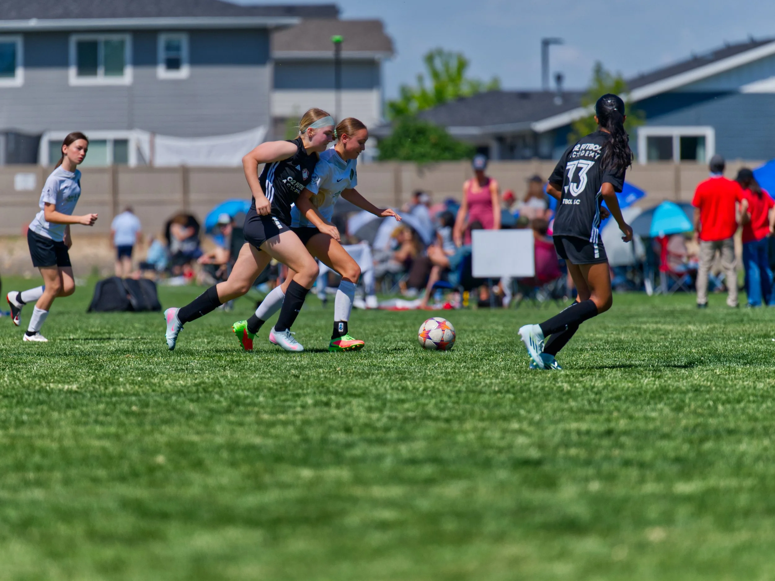 Women playing soccer on a grassy field during daytime, with spectators and houses in the background.