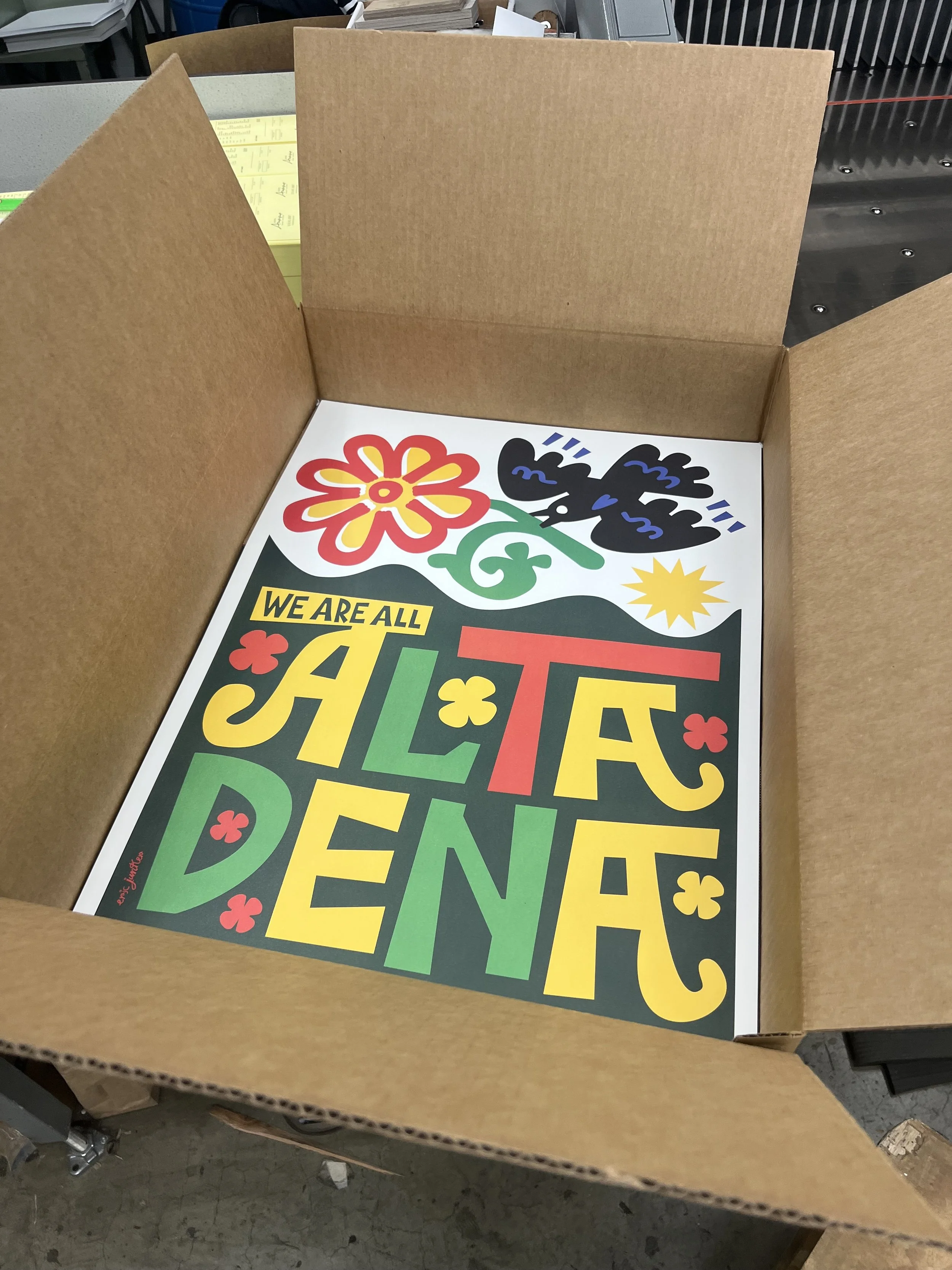 Colorful poster inside a cardboard box featuring the phrase 'We are allALT DENA' with decorative flowers, a butterfly, and a sunrise in bright colors.