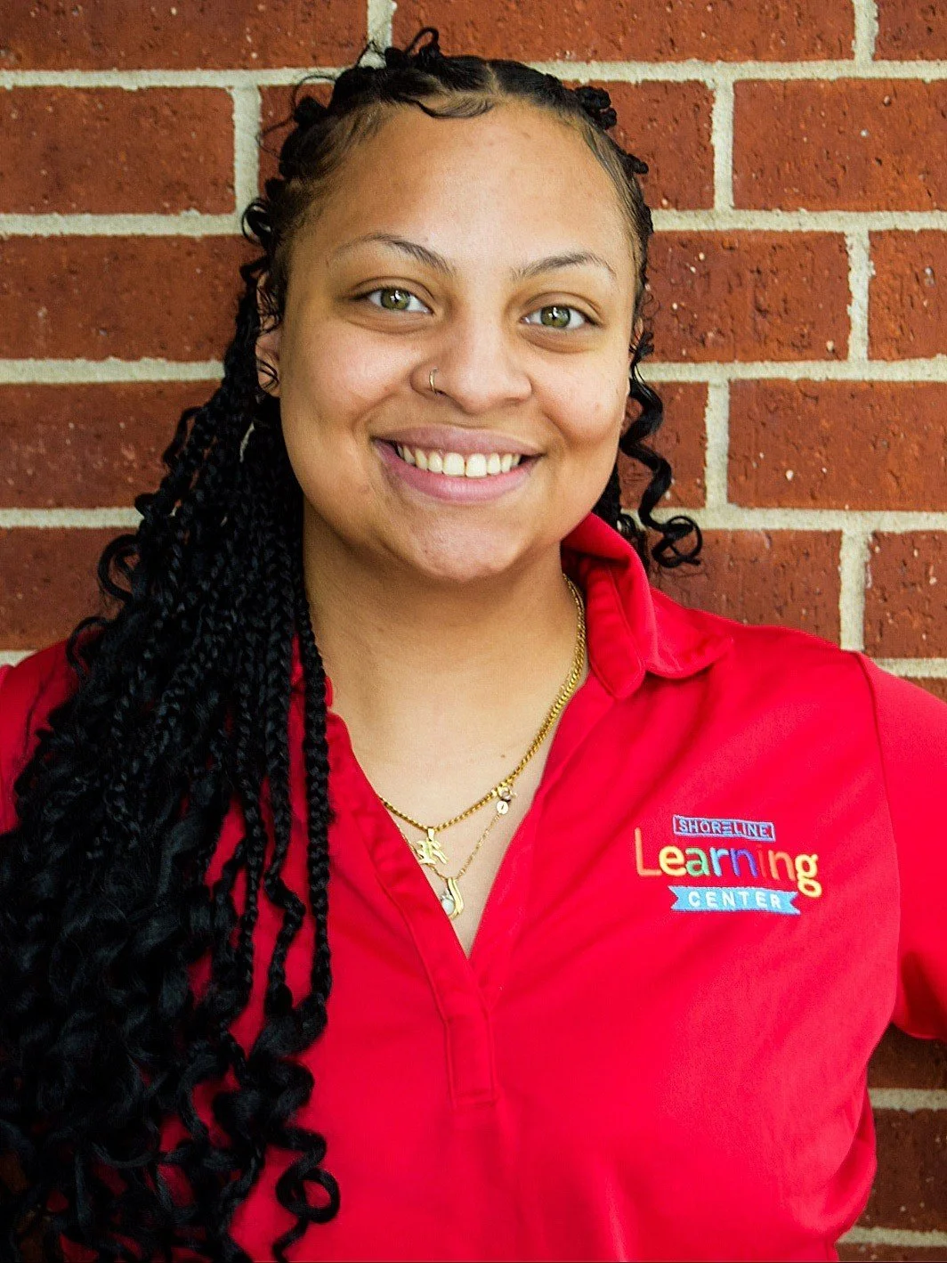 A woman with long curly black hair and light brown skin, smiling, wearing a red jacket with a logo that says 'Shoreline Learning Center,' standing in front of a brick wall.