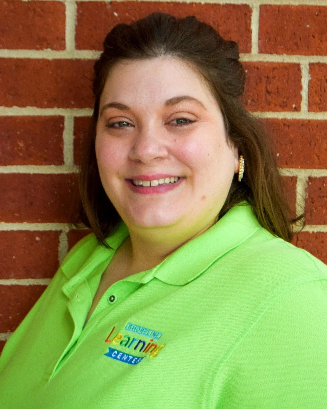 A woman with brown hair and earrings wearing a lime green polo shirt with a logo, standing against a brick wall.