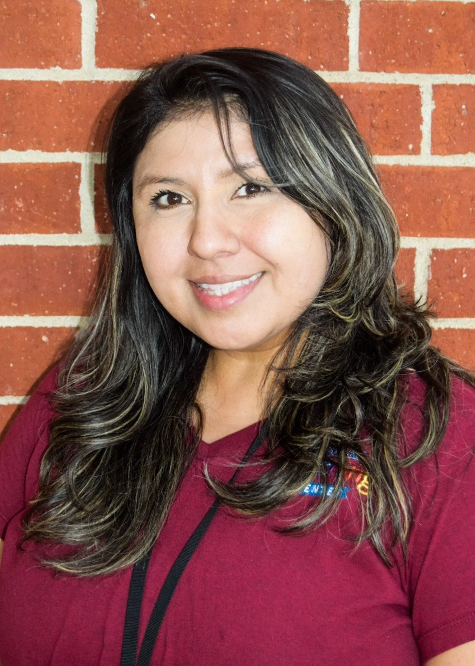 A woman with long dark hair with blonde highlights standing against a red brick wall, smiling at the camera, wearing a maroon shirt.