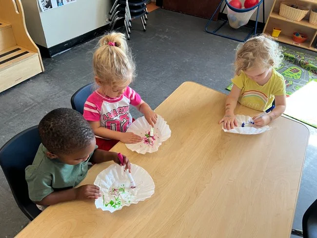 Three young children sitting at a wooden table, decorating paper fans with markers and stickers in a classroom or playroom.