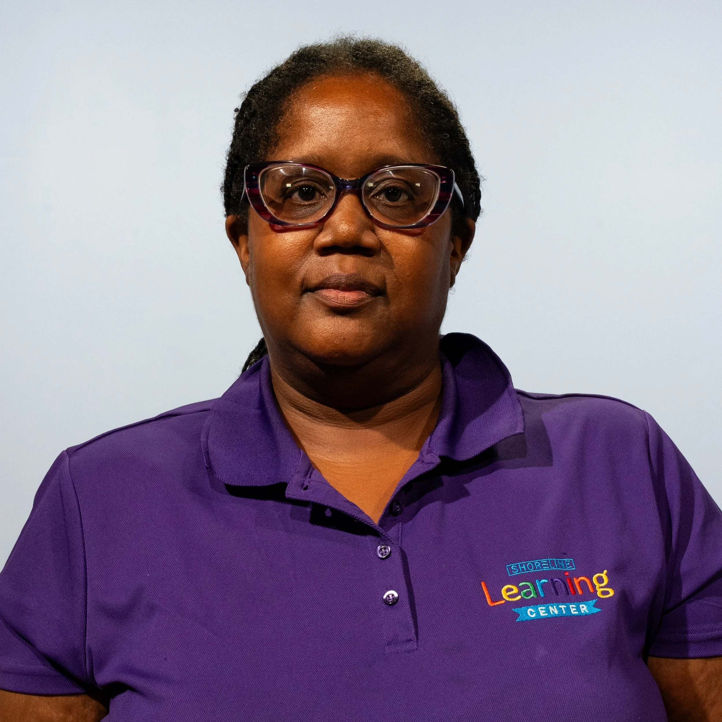 A woman wearing glasses and a purple polo shirt with the logo for Shoreline Learning Center, standing against a plain background.